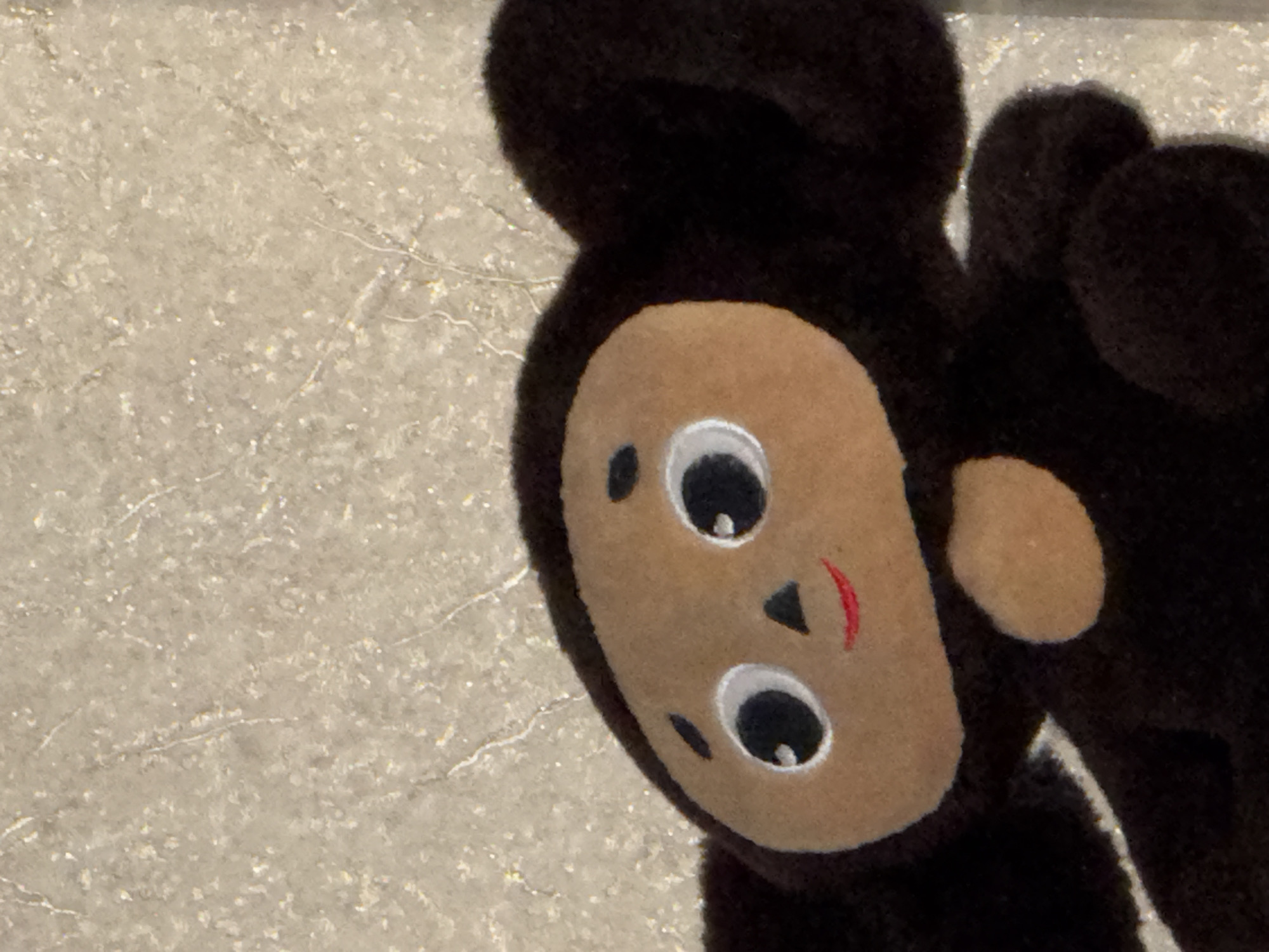 A close-up shot features a dark brown, plush Cheburashka toy positioned to the right of the frame. Its light-brown face, adorned with large, white eyes, a triangular nose, and a red curved smile, is prominent. Part of its ear and body are visible. The background consists of a textured, light-colored surface with subtle patterns, appearing like a stone or concrete slab. While the location is inferred to be Yerevan, Armenia, from context, there is no explicit location information in the image itself. The scene contains no people, actions, or interactions.