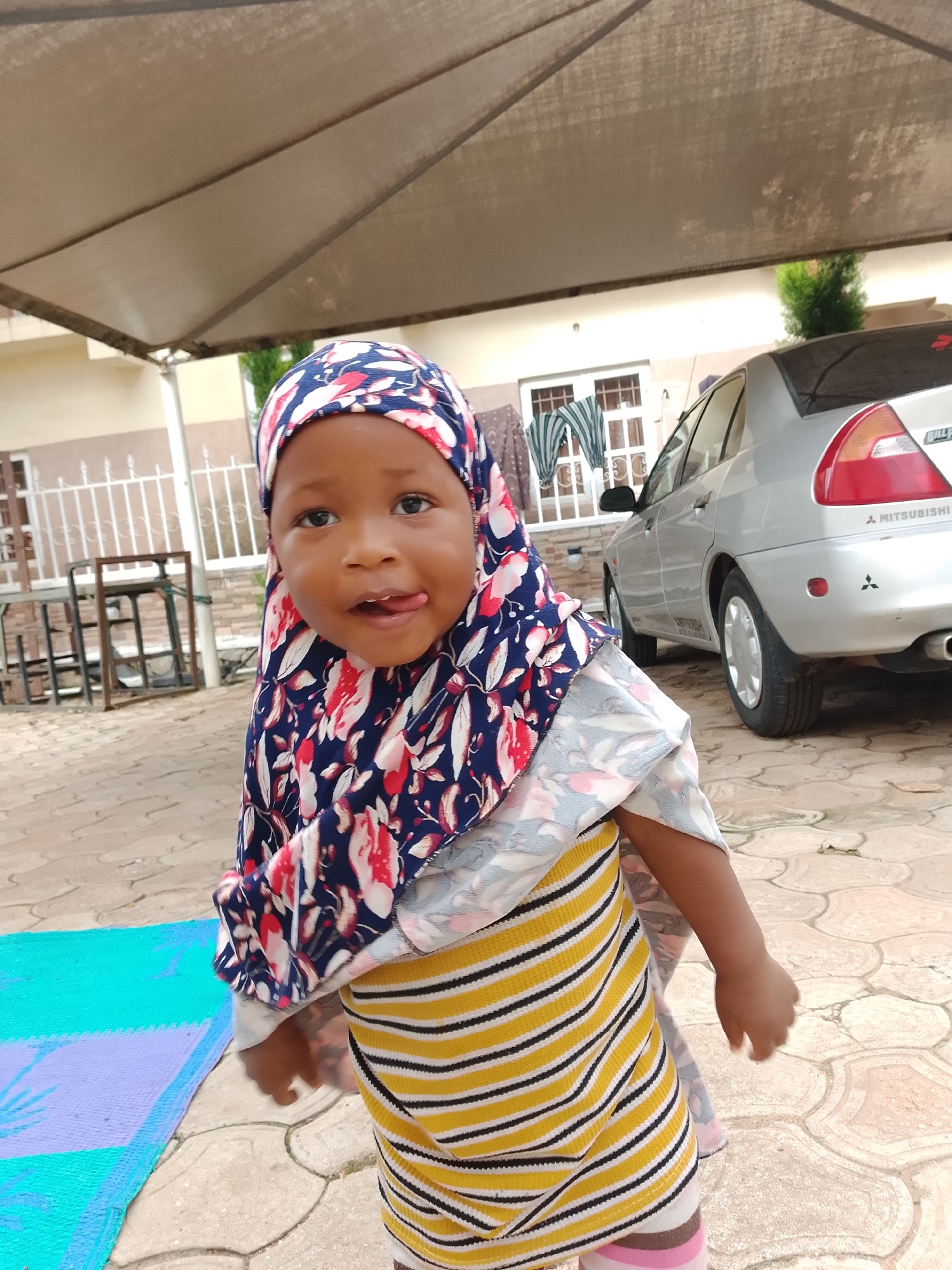 A young child, likely a toddler, is pictured outdoors near a building and a car. The child is wearing a blue and red floral hijab and a yellow and black striped shirt. The child's tongue is slightly extended. The ground is paved with interlocking stones. In the background, a silver Mitsubishi car is parked. A white metal fence and a building with windows are also visible. A light brown colored canopy is overhead. There is a blue and purple mat on the ground to the left of the child. The scene appears to be in a residential area.