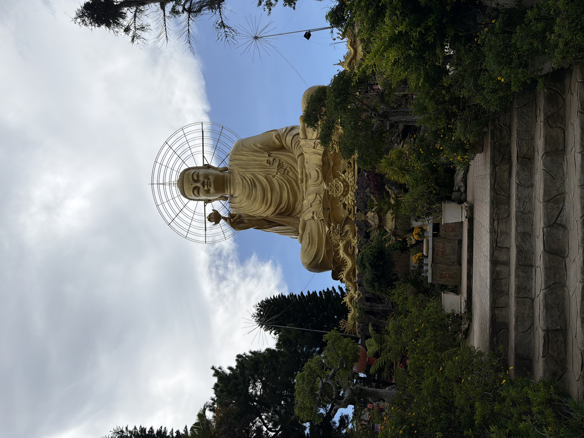 A large, golden Buddha statue is seated outdoors on a mountain slope in Lâm Viên - Đà Lạt, Vietnam. The statue is illuminated by sunlight and set against a backdrop of a cloudy sky. The Buddha is depicted with a serene expression, holding a lotus flower in its right hand. The statue is adorned with a circular halo. In front of the statue, a golden dragon sculpture encircles the base. The scene is surrounded by lush greenery, including trees and bushes, and a stone staircase leads up to the statue.