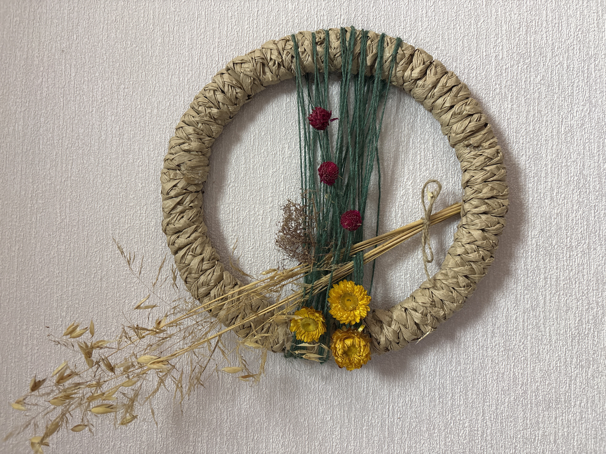 A circular wreath crafted from braided brown paper, approximately 12 inches in diameter, is displayed against a textured white wall. The wreath is adorned with a collection of dried botanical elements. To the left, a cluster of dried grass stalks with seed heads rises from the wreath. Three small, bright yellow dried flowers are also attached to this side. Across the center of the wreath, several strands of dark green yarn are wrapped horizontally. Interspersed with the yarn are three small, dark red dried globe amaranth flowers and a sprig of dried brown foliage. A thin piece of twine is tied in a bow on the lower left portion of the wreath, securing some of the dried stems. The overall impression is a rustic, handmade decorative item. The location is described as Ветрынскі сельскі Савет, Belarus.