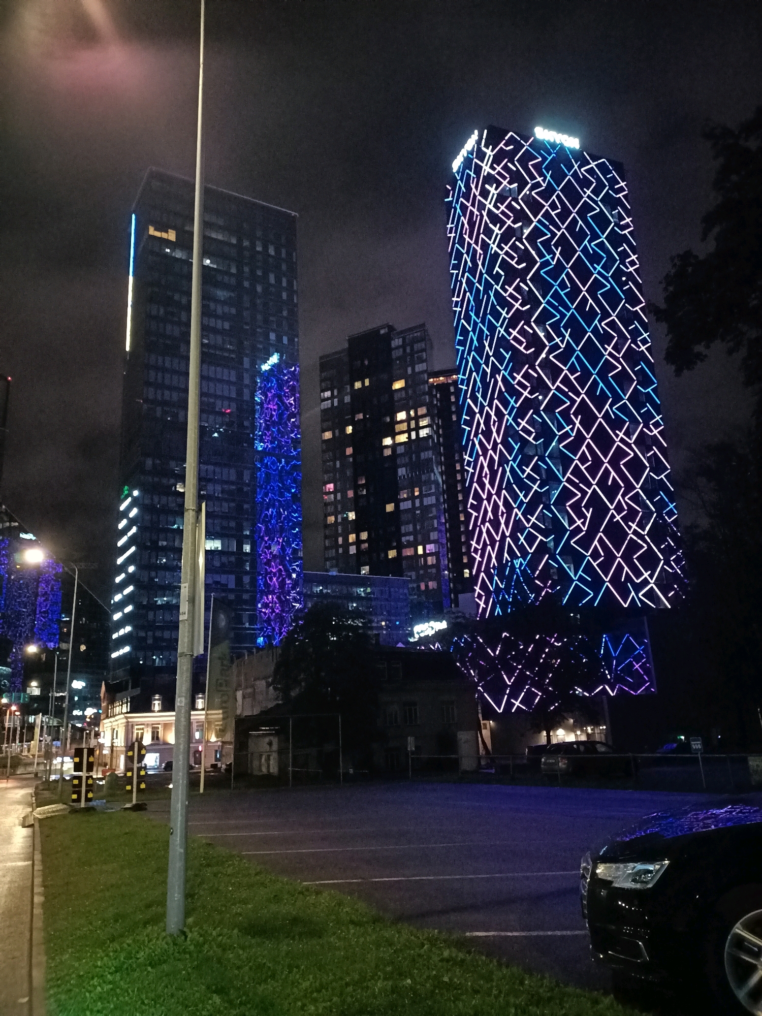 The image captures a nighttime scene in Tallinn, Estonia, dominated by modern skyscrapers illuminated with colorful LED lights. In the foreground, a black Audi car is parked in an asphalt lot, with its front right quarter visible. To the left of the car, a strip of green grass meets the edge of the parking lot. Further into the scene, a tall, slender pole stands vertically in the center. Beyond this pole, several buildings rise into the dark sky. The most striking are two skyscrapers. The one on the right is adorned with a complex geometric pattern of blue and pink lights, creating a vibrant visual display. To its left, another dark skyscraper features vertical white LED lights along its edge and a pattern of blue and purple lights on another section. In the background, other buildings with illuminated windows are visible, suggesting an urban environment. The sky is dark and appears slightly hazy, possibly due to mist or clouds. The overall impression is one of a contemporary cityscape at night, with a focus on architectural lighting.