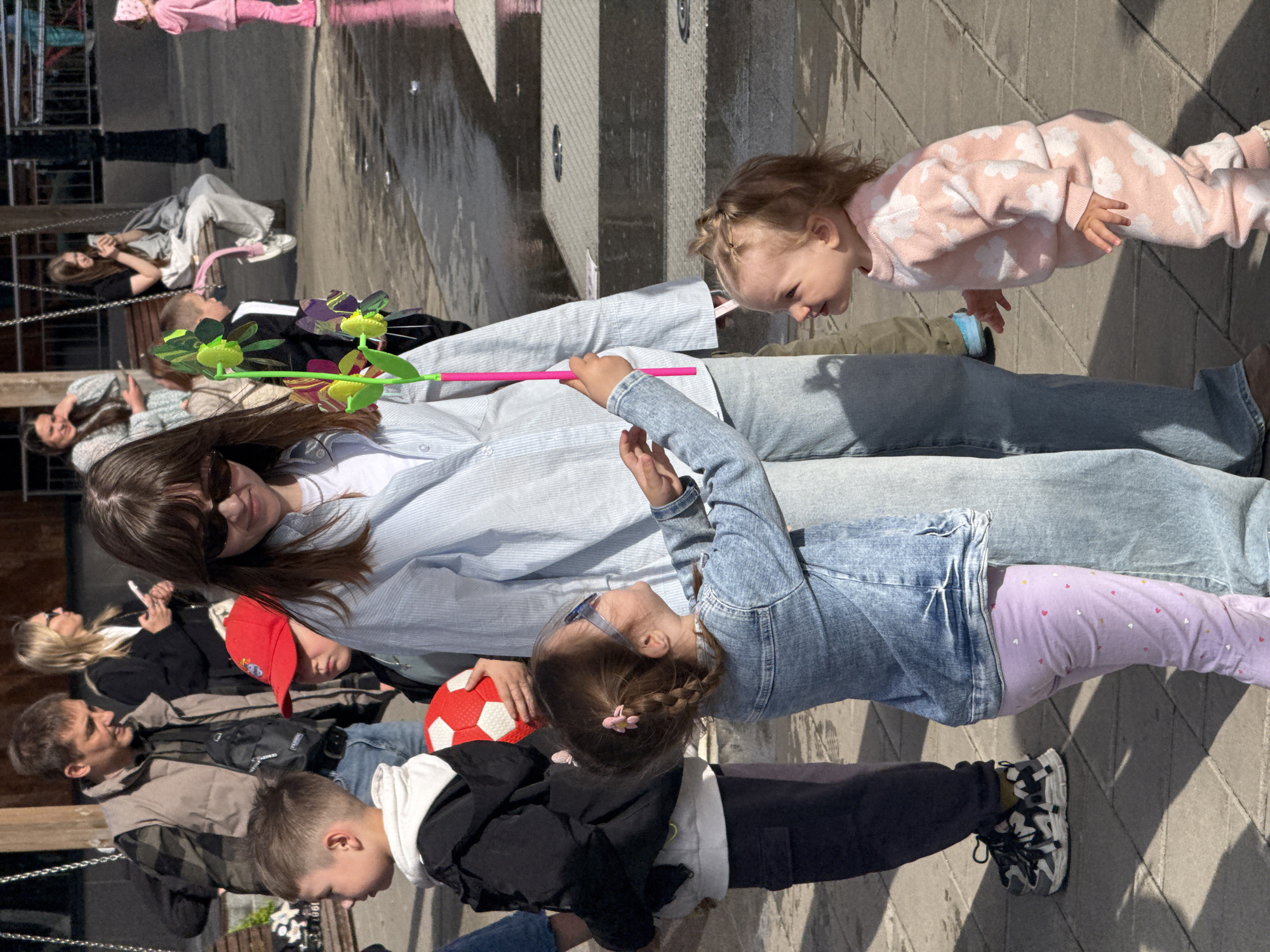 The image shows a group of people, including children and adults, likely in a public space. The subjects are arranged in a way that suggests they are interacting or posing for a photo. A woman wearing sunglasses is lying on the ground, with children positioned on and around her. One child holds a toy pinwheel, another holds a red and white ball. The background features what appears to be a modern water feature or fountain, constructed with light-colored stone. Other people can be seen in the background, some sitting and some standing. The lighting suggests it is daytime. The location is Yekaterinburg, Russia.