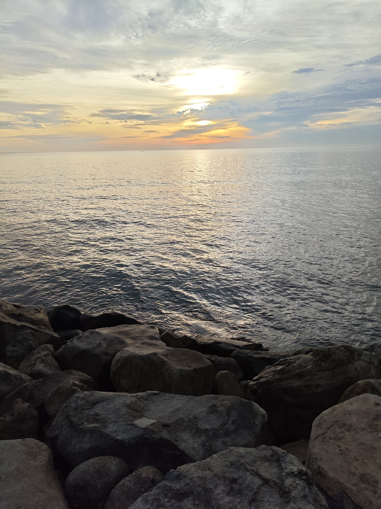 The image depicts a coastal scene at either sunrise or sunset, viewed from a rocky shoreline in Lhokseumawe, Indonesia. In the foreground, numerous large, irregular, dark grey rocks form a prominent mass, extending across the bottom fifth of the frame and receding into the lower corners. These rocks appear to be part of a natural or man-made breakwater. Immediately beyond the rocks, the calm surface of the sea stretches across the midground and background, exhibiting gentle ripples that reflect the ambient light. The water transitions from darker tones near the shore to lighter, shimmering golden hues where the sun's reflection is most intense. The horizon line is visible, separating the sea from the sky. The sky, occupying approximately the upper two-thirds of the image, displays a gradient of colors characteristic of a crepuscular period. A bright, diffused light source, indicative of the sun, is positioned low above the horizon towards the center-right, casting a warm yellow-orange glow. This glow blends into lighter yellow, orange, and pale grey-blue tones across the expansive sky, which is interspersed with thin, wispy cloud formations. No individuals, vessels, or specific man-made structures are discernible within the frame. The scene conveys a tranquil natural environment.