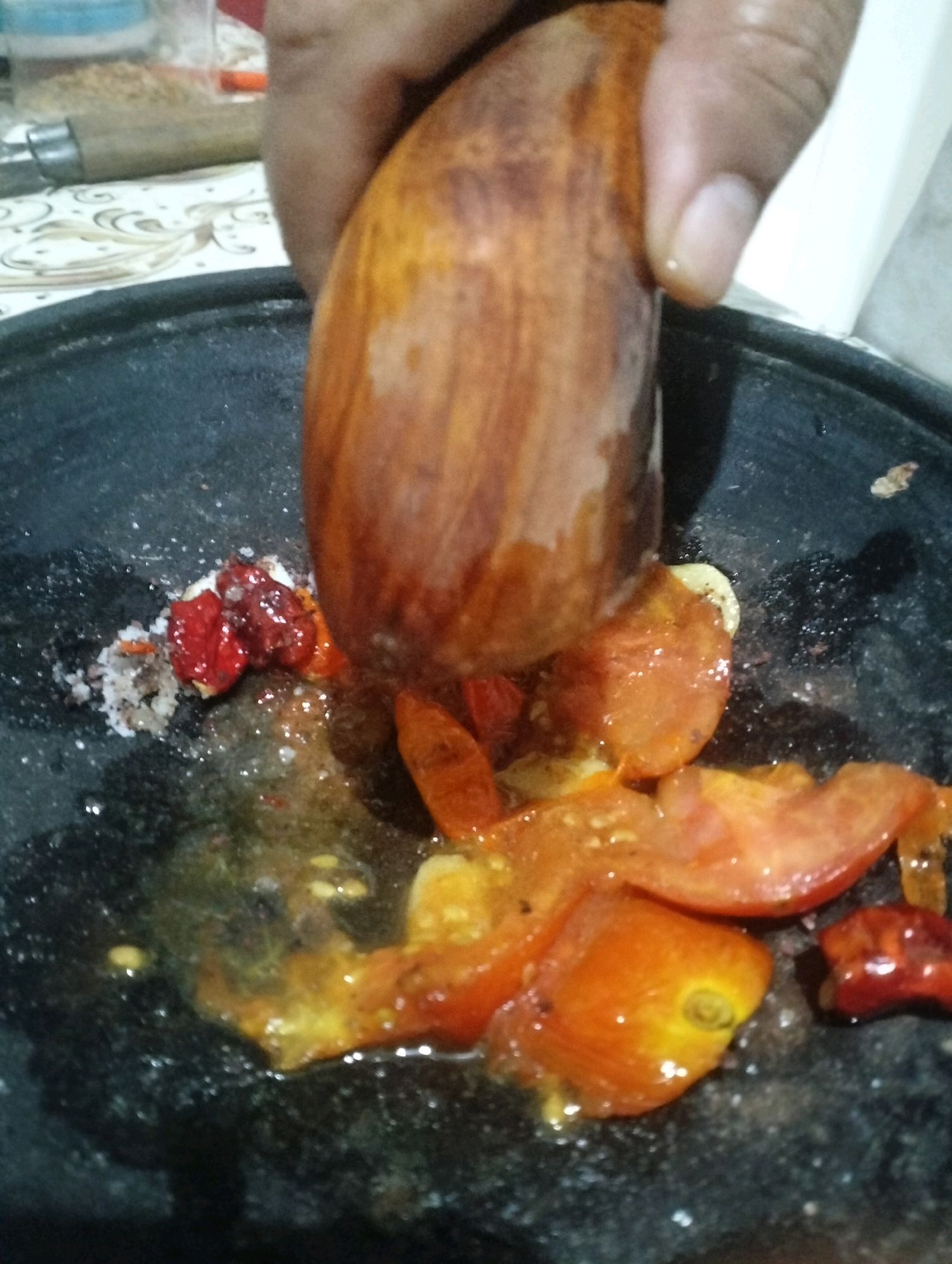 A person's hand is holding a wooden pestle over a dark, circular mortar containing chopped tomatoes, chili peppers, and salt. The mortar is positioned on a patterned surface. The scene appears to be an indoor setting in Klecorejo, Indonesia, likely in a kitchen, where ingredients are being prepared.