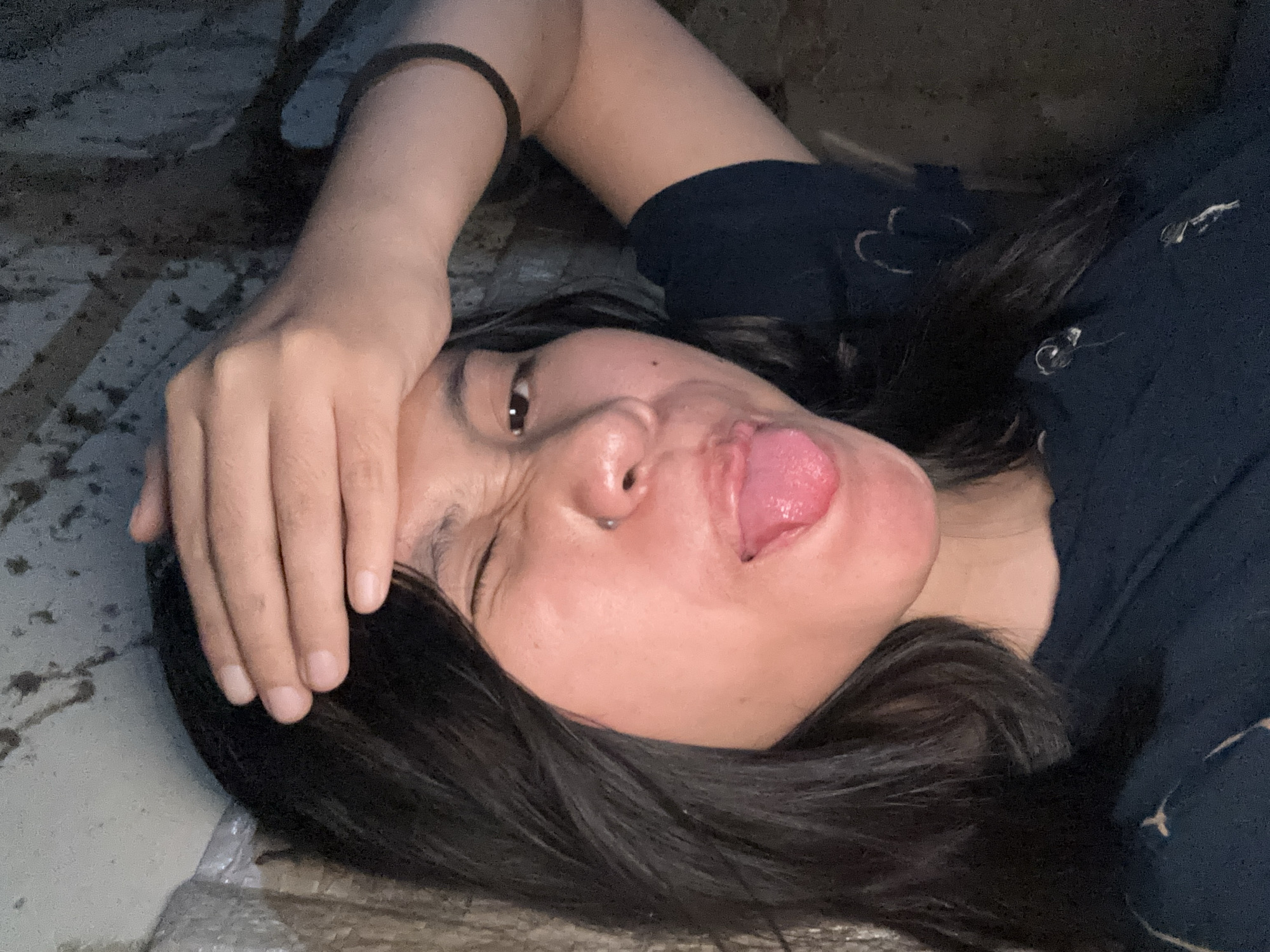 A young woman with dark hair is shown from the chest up, lying down. She is touching her forehead with her right hand, and her left eye is closed in a wink. Her tongue is sticking out and is bright pink. She is wearing a dark blue t-shirt. The background appears to be a rough, textured wall with some dirt or debris on it. There is also a piece of what looks like plastic sheeting or tarpaulin on the left side of the image. The lighting is dim, creating shadows and highlighting the woman's features. The scene is likely indoors, possibly in a utility area or an unfinished space within a building in East Jakarta, Indonesia.