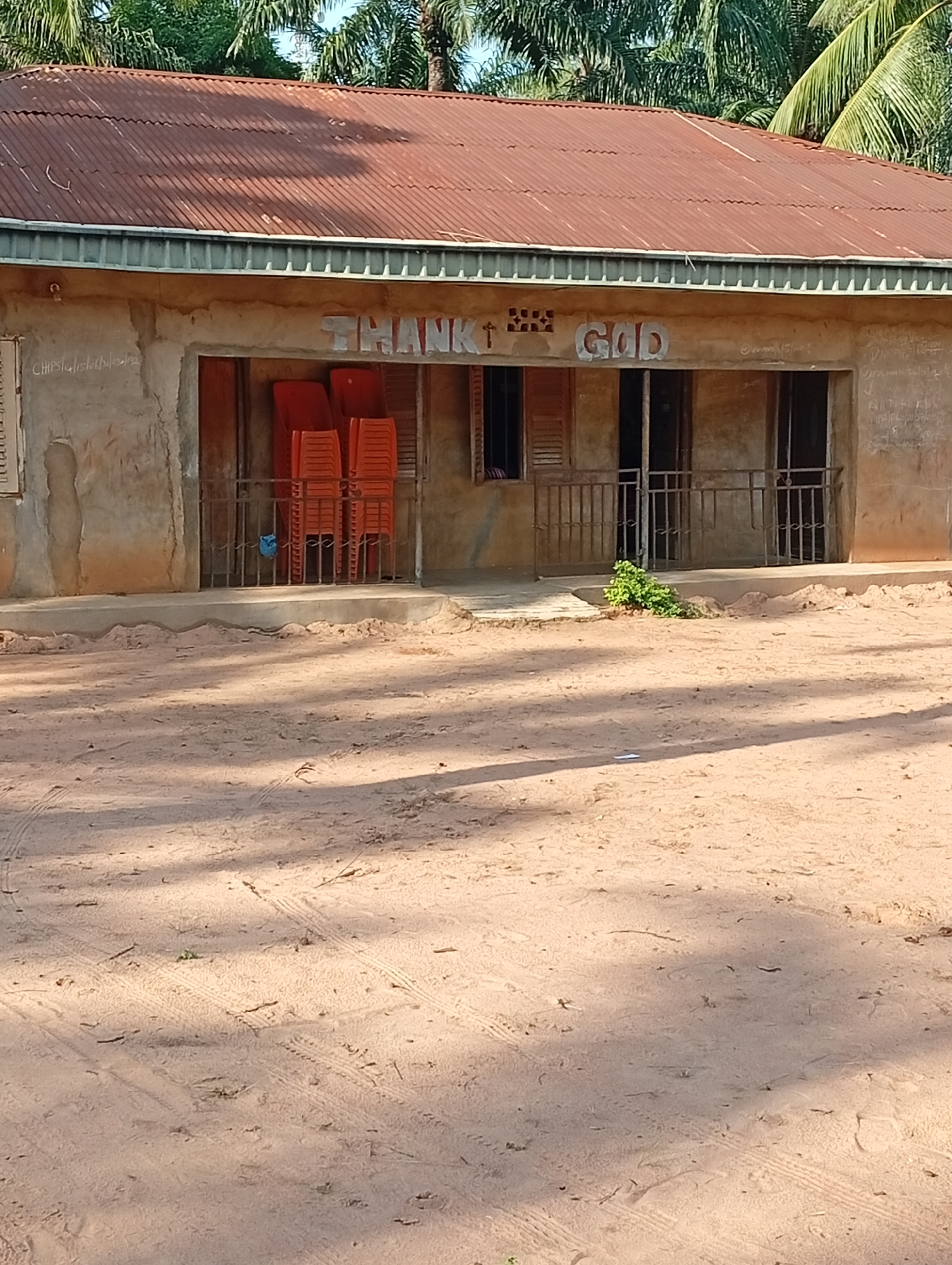 The image depicts the exterior of a building in Adda, Nigeria, with a red corrugated metal roof. The building's walls are made of a rough, plastered material. White, raised lettering on the facade reads "THANK GOD". There are multiple openings, some with windows and others that appear to be doorways. In front of the building, a metal railing encloses an area where several stacks of bright orange plastic chairs are visible. A small, green bush is growing near the railing. The ground in front of the building is sandy and appears to be a dirt road, with tire tracks visible. The overall scene suggests an outdoor setting, possibly a community center or a place of worship.