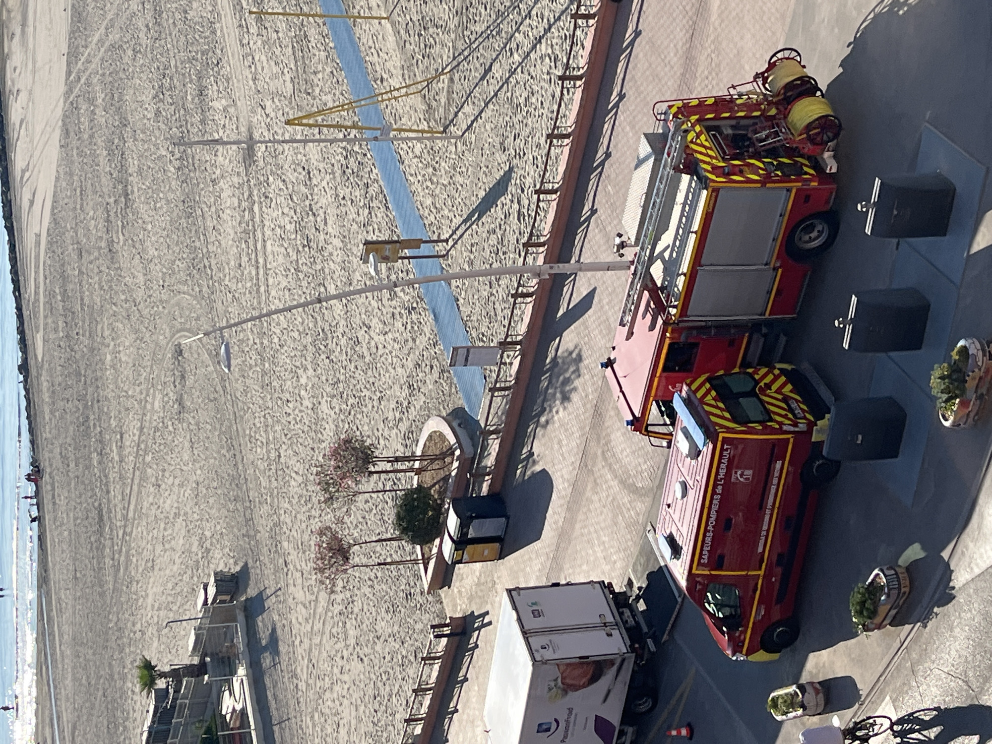 The image shows a view from an elevated position looking down at a sandy beach and a paved area next to it. In the paved area, there are two fire trucks: a red van with "Sapeurs-Pompiers de l'Herault" written on its side and a larger red fire engine with equipment visible. To the left of the fire trucks, a white refrigerated truck is parked. In the background, the sea is visible with a few people on the beach and in the water. The scene is likely in Palavas-les-Flots, France, given the presence of "Herault" on the fire truck, as Herault is a department in the Occitanie region of France where Palavas-les-Flots is located. The environment is a public area, possibly a coastal town, with elements of both public services (fire trucks) and recreational activity (beach).