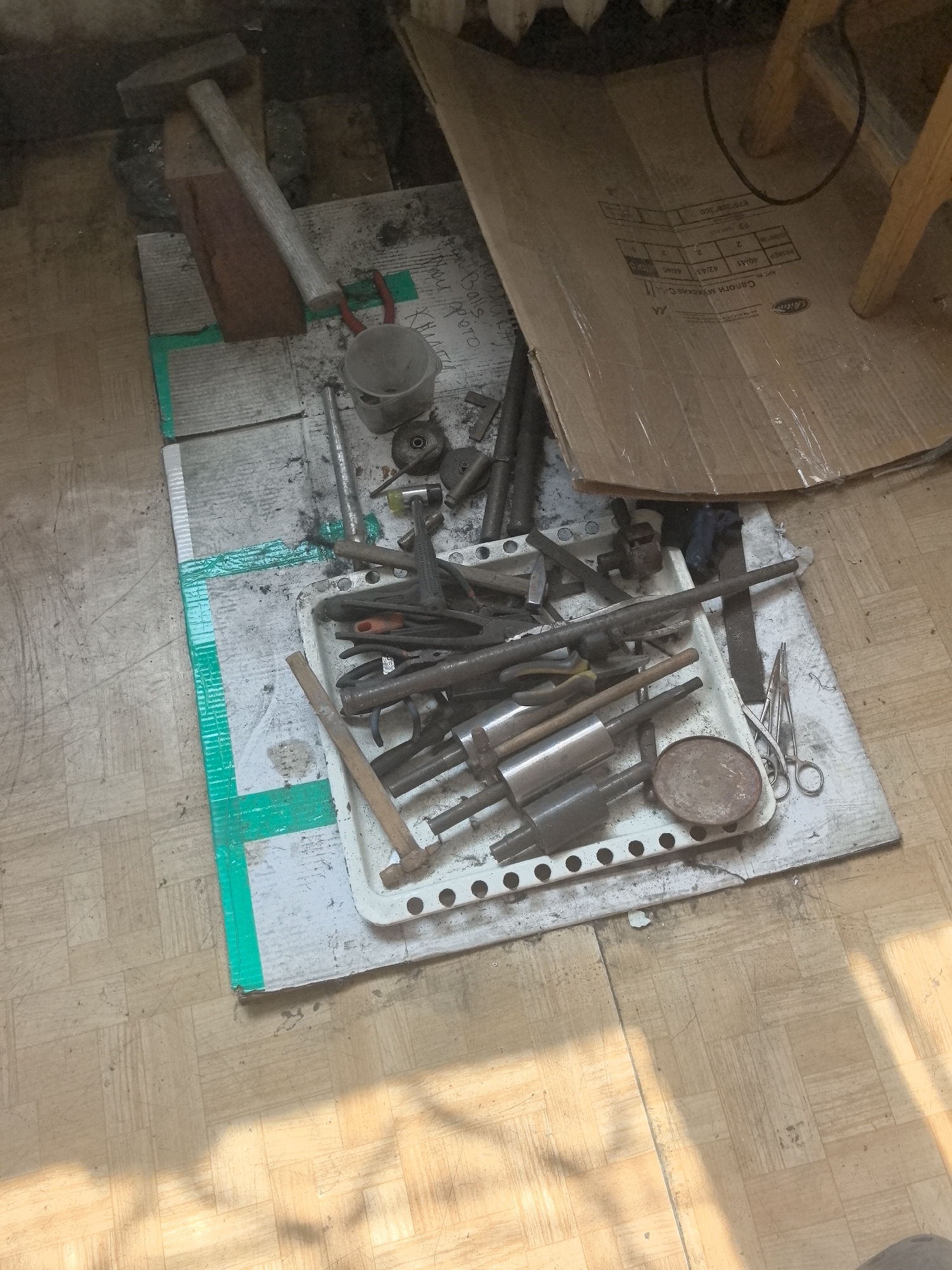 The image depicts a cluttered workspace on a light-colored, tiled floor, likely indoors. The scene is dominated by various tools laid out on a white tray and scattered around it. To the left, a large hammer rests on a piece of wood, which is itself placed on a surface marked with green tape. Several metal rods and cylindrical objects are visible within the tray, alongside pliers, a small circular metal object, and a pair of silver scissors. A cardboard box lies partially opened in the upper right corner, propped up by wooden legs of what appears to be a table or stool. The floor shows signs of wear and tear, with scuff marks and dust present. Sunlight casts long shadows across the floor, indicating a bright day. The overall impression is one of a DIY or repair area. Without additional context, pinpointing the exact location within Amursk, Russia, is impossible, but the scene suggests a private workshop or storage space.