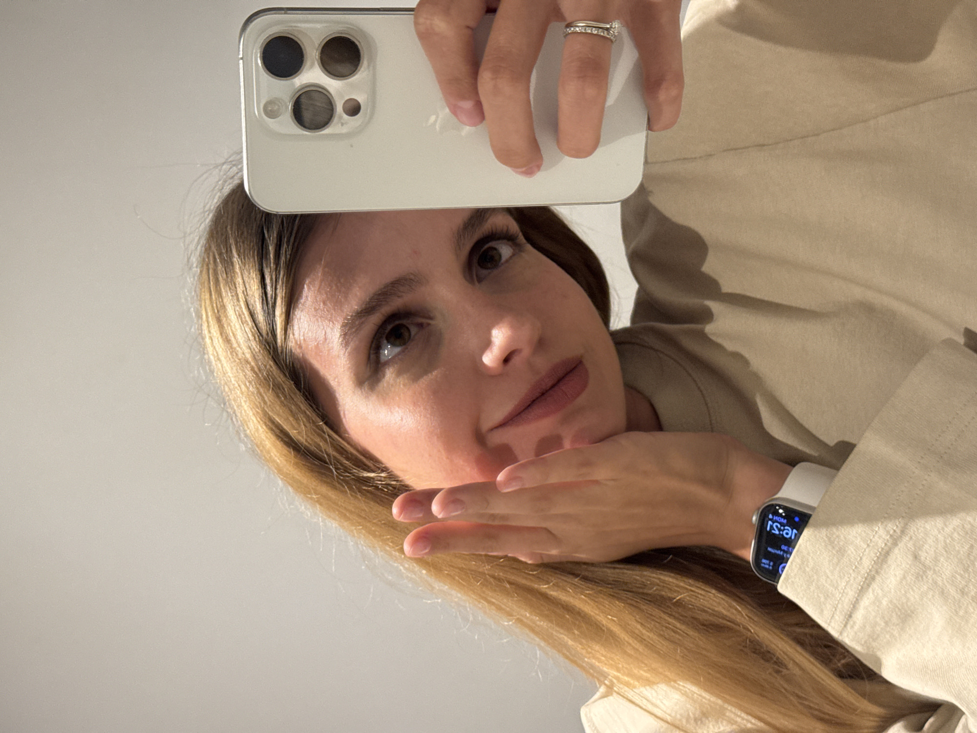 A woman with long blonde hair is taking a selfie with a white smartphone. She is wearing a tan-colored jacket and a white smartwatch is visible on her wrist. The location is unknown.