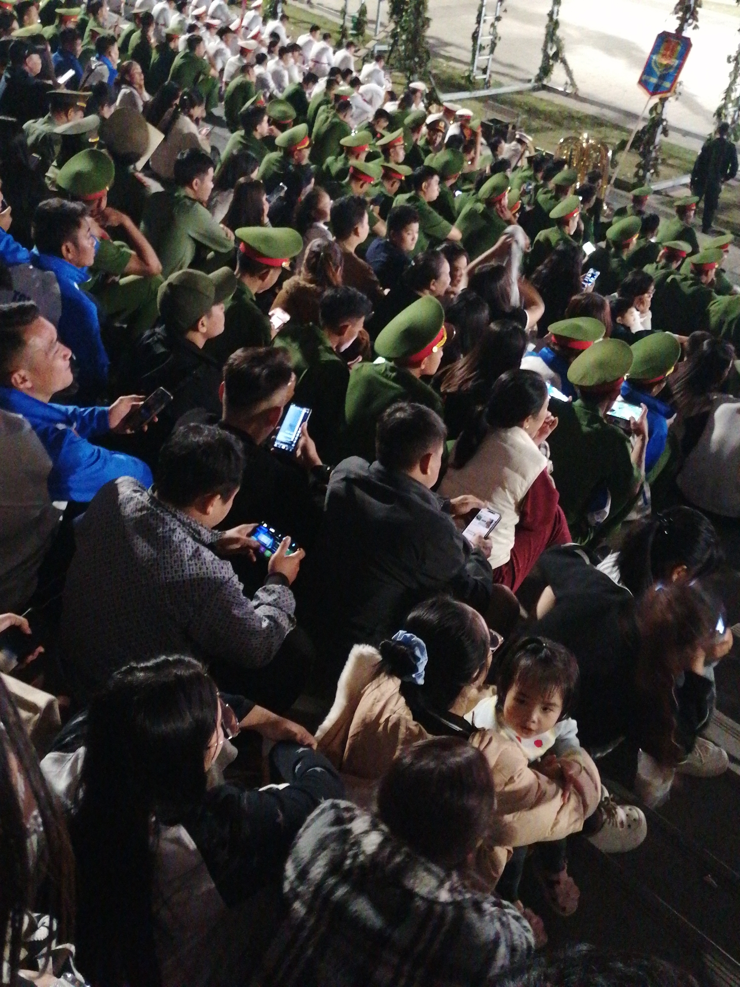 The image depicts a large crowd of people, predominantly young adults and children, gathered in an outdoor setting at night. Many individuals are wearing green uniforms and hats, suggesting they may be military personnel or cadets. The lighting is artificial, casting a warm glow and creating shadows, indicating it is nighttime. Some people are holding up their mobile phones, possibly to take pictures or videos of an event, or perhaps using them for entertainment.
In the foreground, a young child is being held by an adult, both looking towards something outside the frame. The child is wearing a light-colored jacket, and the adult is wearing a beige vest over a maroon garment. Other individuals in the crowd are seen in various states of engagement: some are looking intently at their phones, while others appear to be observing something in front of them.
The atmosphere seems to be one of a public gathering or event, possibly a parade, festival, or military ceremony, given the uniformed individuals and the excited crowd. The presence of what looks like a ceremonial banner or flag at the top right of the image further supports this idea. The overall impression is one of a lively, engaged crowd participating in a nighttime event in a public space, possibly in Da Lat, Vietnam. No visible text is present in the image.