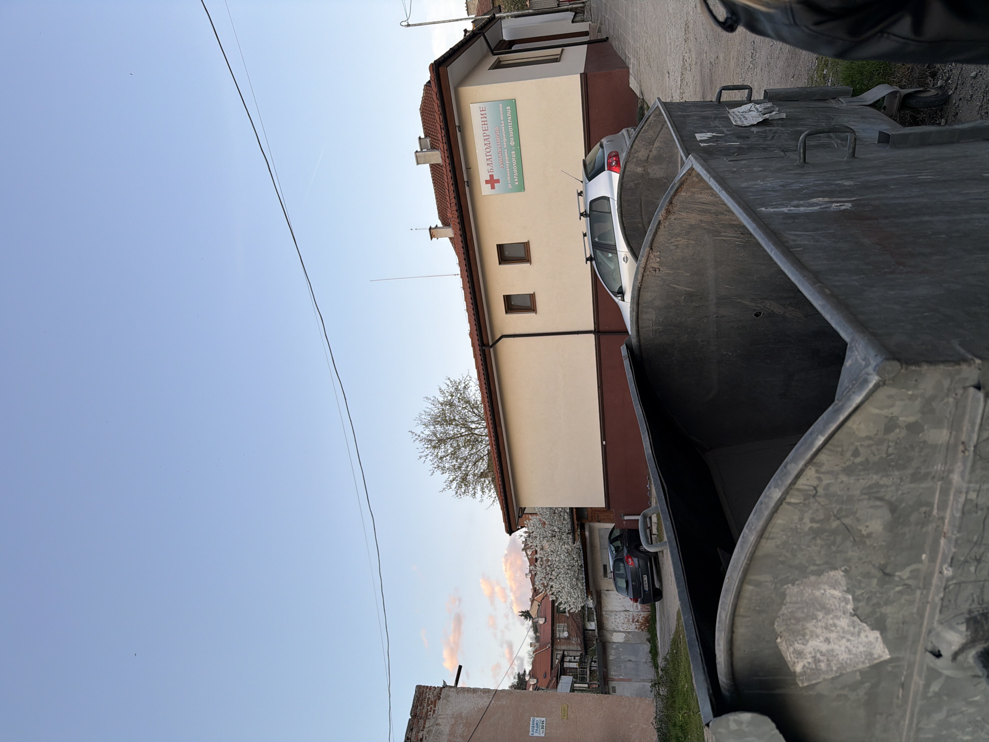 The image, taken in Panagyurishte, Bulgaria, shows an outdoor scene. A large, dark gray metal dumpster dominates the foreground, obscuring portions of the background. Behind the dumpster, a beige and brown two-story building with a red tile roof is visible. A sign on the building reads "БЛАГОДАРЕНИЕ ЗА ПЪРВИЧНИ МЕДИЦИНСКИ ПРЕГЛЕДИ, КОНСУЛТАЦИИ, КАРДИОЛОГИЯ, ФИЗИОТЕРАПИЯ," indicating medical services. A silver car is parked near the building. In the background, a row of residential buildings with red tile roofs is seen, interspersed with blooming trees. A blue sky with light clouds is in the upper left corner. Power lines are visible across the sky. Another dark-colored car is parked behind the silver car. The image appears to be taken from a low angle, possibly at street level.