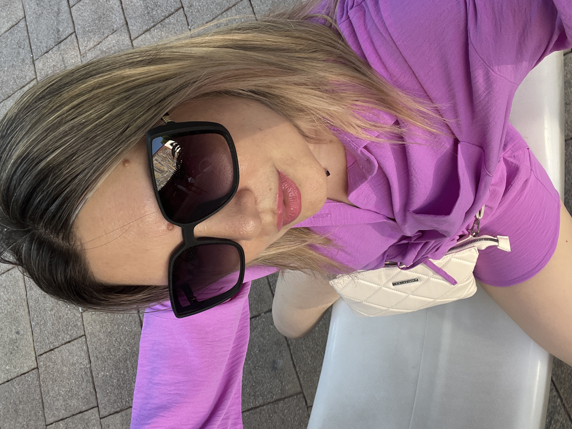 A person is posing for a selfie outdoors, lying down and looking upwards towards the camera. They are wearing a vibrant purple top and black-rimmed sunglasses. A small, white quilted purse with a silver zipper and a purple strap detail is visible on their lap. The background consists of grey paving stones and what appears to be a white bench or surface. The lighting suggests it is daytime, likely with sunlight. The person's hair is blonde with darker roots and is casually styled, partially obscuring their face and shoulder. The overall impression is one of leisure and personal style. There is no visible text.