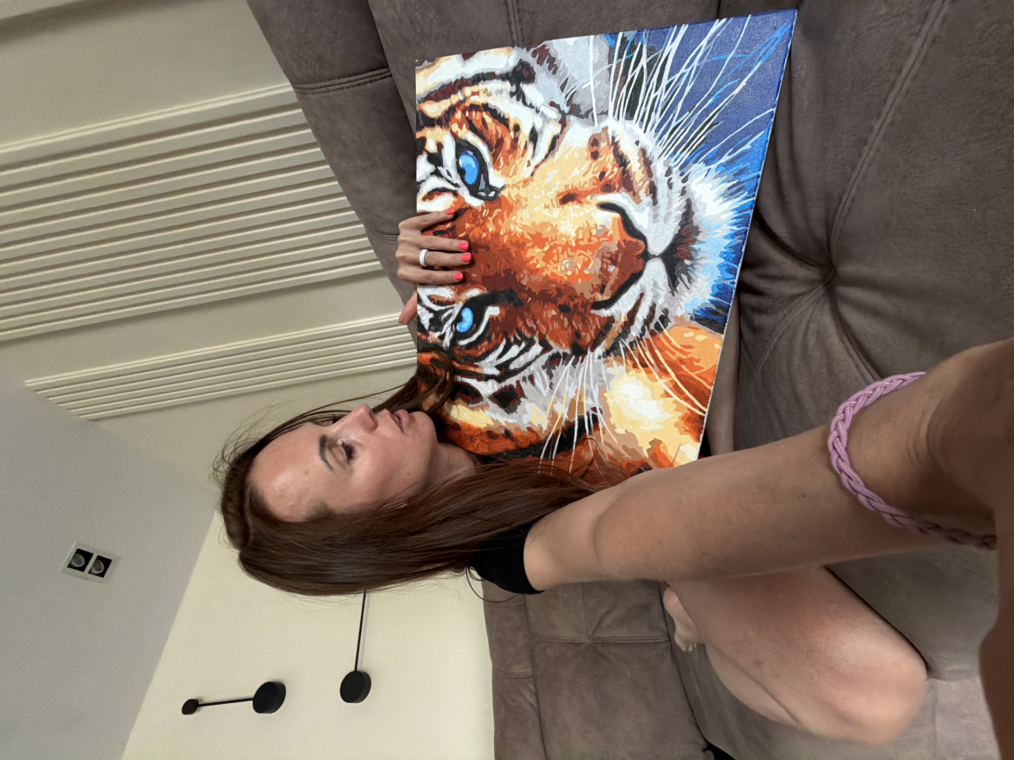 A young woman is captured indoors, likely in a home setting in Birobidzhan, Russia, holding up a vibrant painting of a tiger's head. She has long, straight brown hair and is wearing a black top. Her left hand, featuring bright pink nail polish and a white ring, supports the painting, while her right arm, adorned with a braided lavender bracelet, extends towards the camera. The woman appears calm and focused, looking towards the artwork she holds.
The painting is a colorful, stylized depiction of a tiger's face, characterized by bold orange, white, and black stripes, with piercing blue eyes as a central feature. The background of the painting is a mix of blue and white, with white lines radiating outwards, possibly representing whiskers or an abstract artistic effect. The distinct color blocking suggests it may be a paint-by-numbers piece.
The setting includes a brown upholstered couch, a light-colored wall with horizontal ridged paneling, and a plain white wall. On the white wall, there's a modern black decorative object with two circular elements connected by a rod, and a rectangular light fixture with two square lights. The overall lighting suggests it is daytime. There is no visible text in the image.