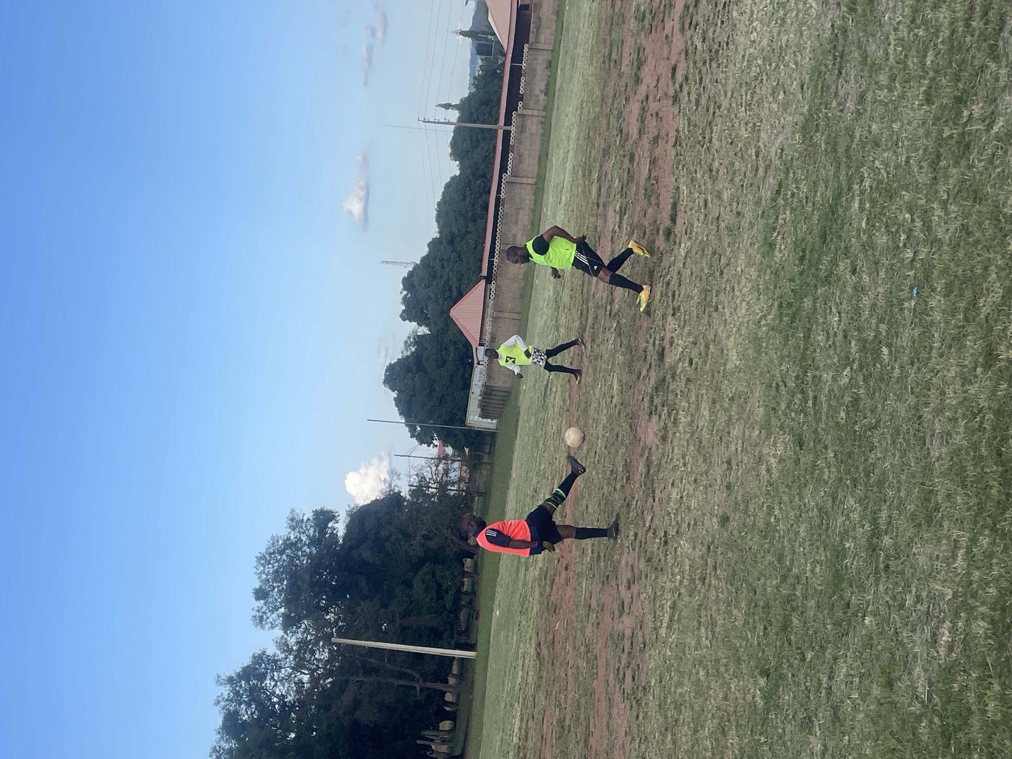 Three individuals are on a grassy field, presumably a sports ground, under a clear blue sky. The ground is uneven and patchy, with areas of dry earth and sparse grass. In the background, there is a row of trees, a fence with a repetitive pattern, and some buildings with reddish roofs. A few distant structures and power lines are also visible.
On the left, a person wearing an orange vest, black shorts, and black socks with a green stripe is kicking a soccer ball. They are positioned with their left leg extended towards the ball, mid-kick. To the right of this person and slightly further back, another individual in a white long-sleeved shirt with a yellow logo and patterned shorts is walking. Further to the right and closer to the foreground, a third person in a neon yellow vest, black shorts, and black socks with yellow accents is running. The soccer ball is on the ground between the first two individuals.
The scene appears to be in Jos, Nigeria, suggested by the context provided by the prompt. The lighting indicates it is daytime, possibly late afternoon due to the soft light and long shadows cast by the trees in the background. The overall environment suggests an outdoor recreational or training area.