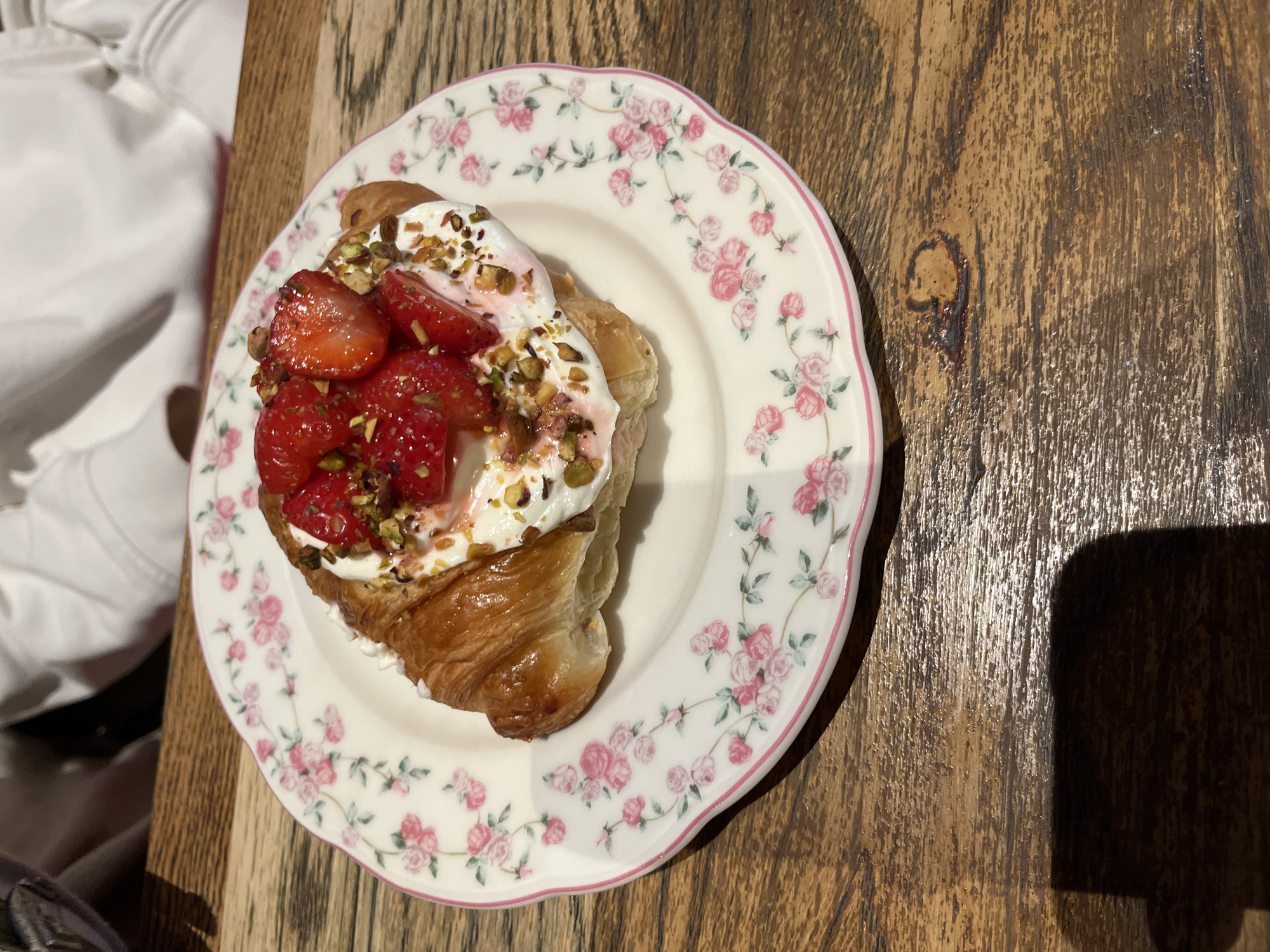 An overhead shot shows a croissant topped with whipped cream, chopped strawberries, and pistachios, resting on a floral patterned plate. The plate is placed on a wooden table, casting a shadow to the left. The background shows the blurred white sleeve of a person. The scene is set at the restaurant "U Andreya" in Svetlogorsk, Russia.