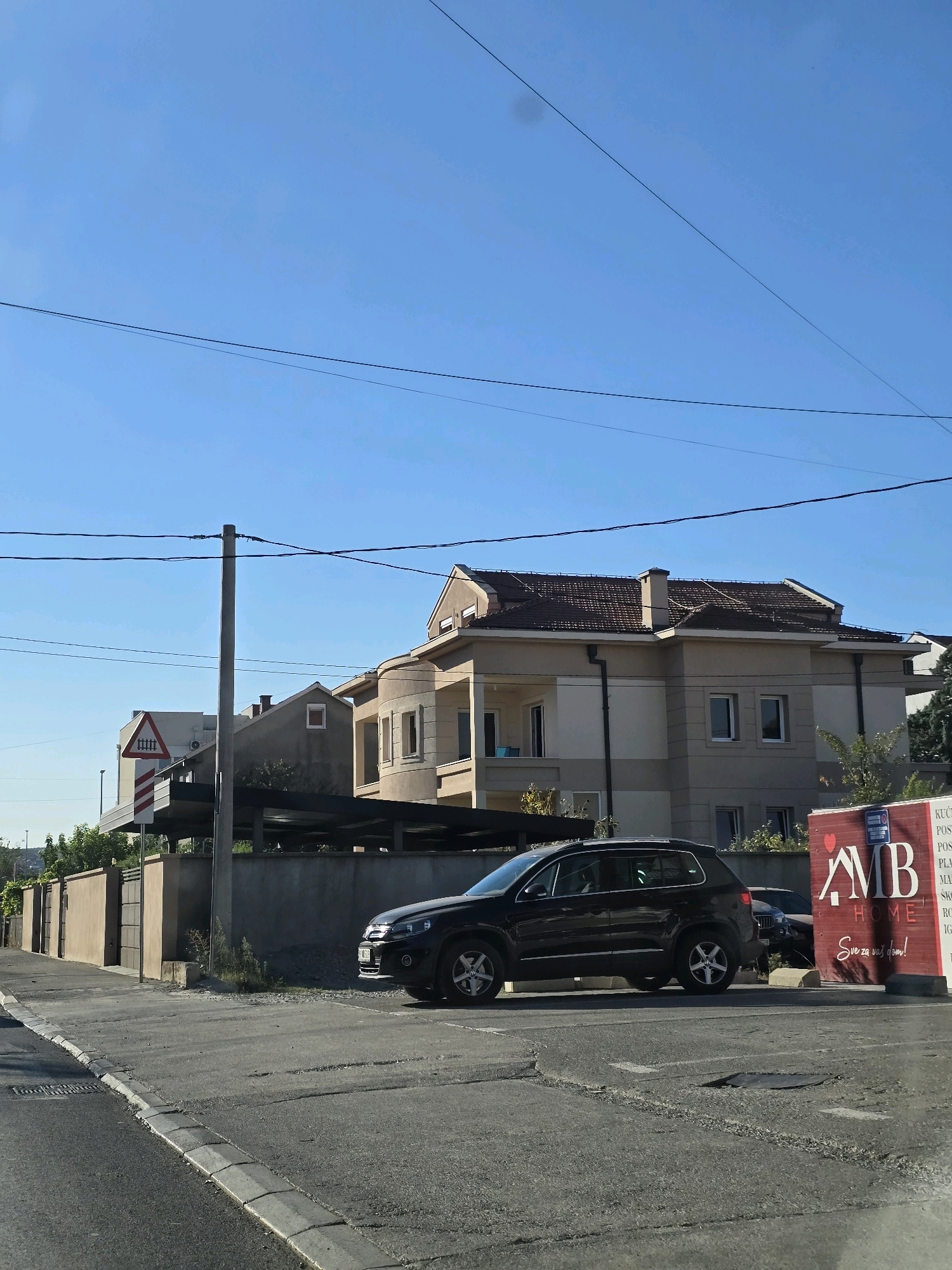 The image shows a street scene in Podgorica, Montenegro. The foreground features a cracked asphalt road and sidewalk. In the midground, a dark-colored SUV is parked on the side of the road. Behind the SUV, a concrete wall separates the road from a residential area. A large, modern house with a tiled roof is visible behind the wall. The house has multiple windows and balconies. A red advertisement sign with the text "AMB HOME" is partially visible on the right side of the image. In the background, there are more houses and trees under a clear blue sky with visible power lines. A traffic sign indicating a railway crossing is located on the left side of the image.
