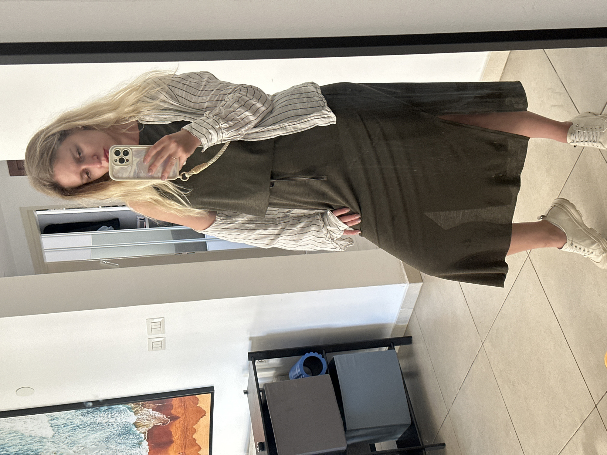 A woman with long blonde hair is taking a mirror selfie. She is wearing a green dress and a striped shirt, and white sneakers. The location is likely indoors, possibly a home in Petah Tikva, Israel. Background elements include a cabinet, a TV, wall outlets, and storage shelves.