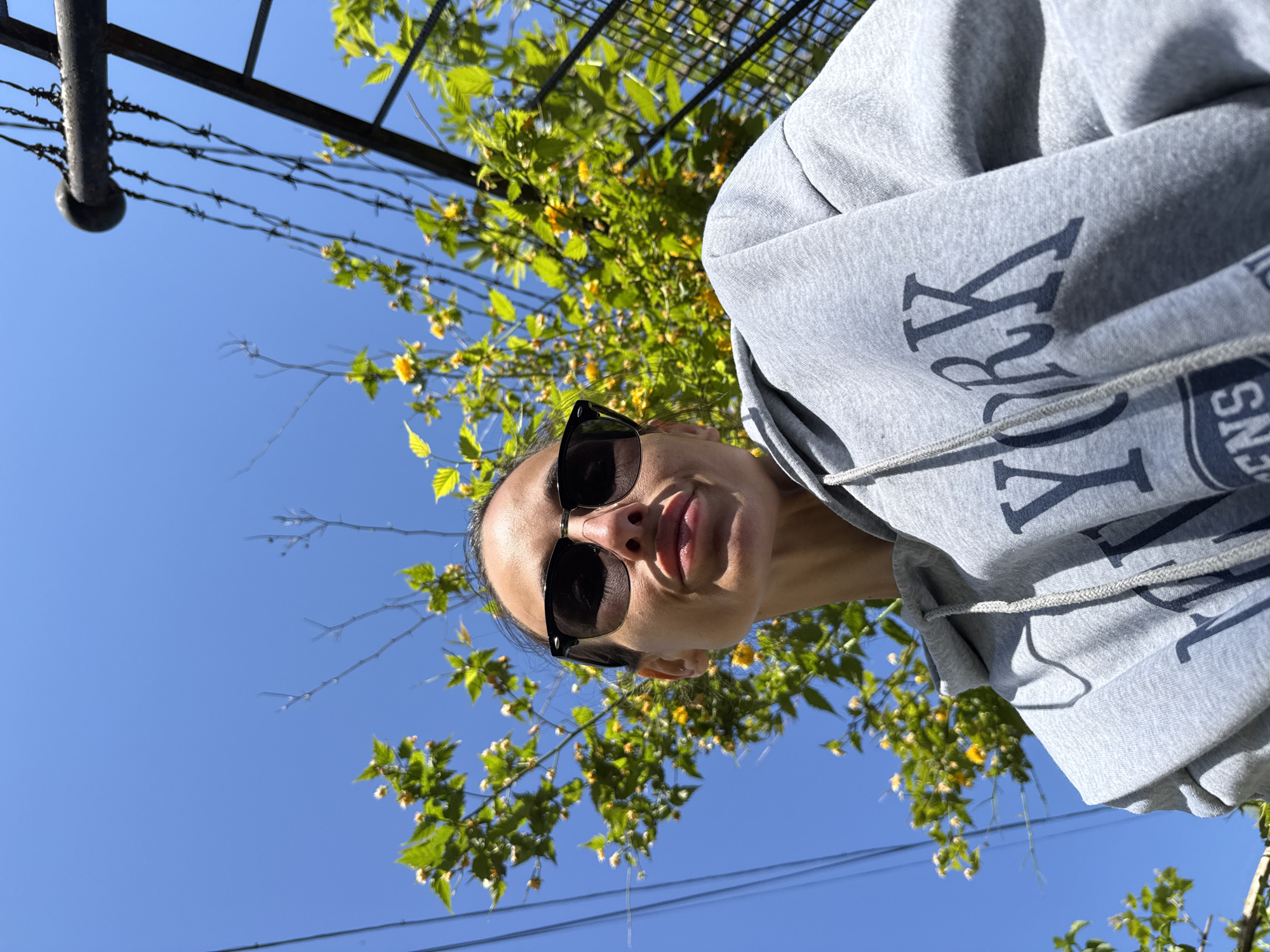 A woman wearing sunglasses and a gray hoodie with "NEW YORK" printed on it is pictured from a low angle against a clear blue sky. Leaves and yellow flowers are visible in the background. There's also a metallic structure with barbed wire. The location is likely Pleven, Bulgaria.