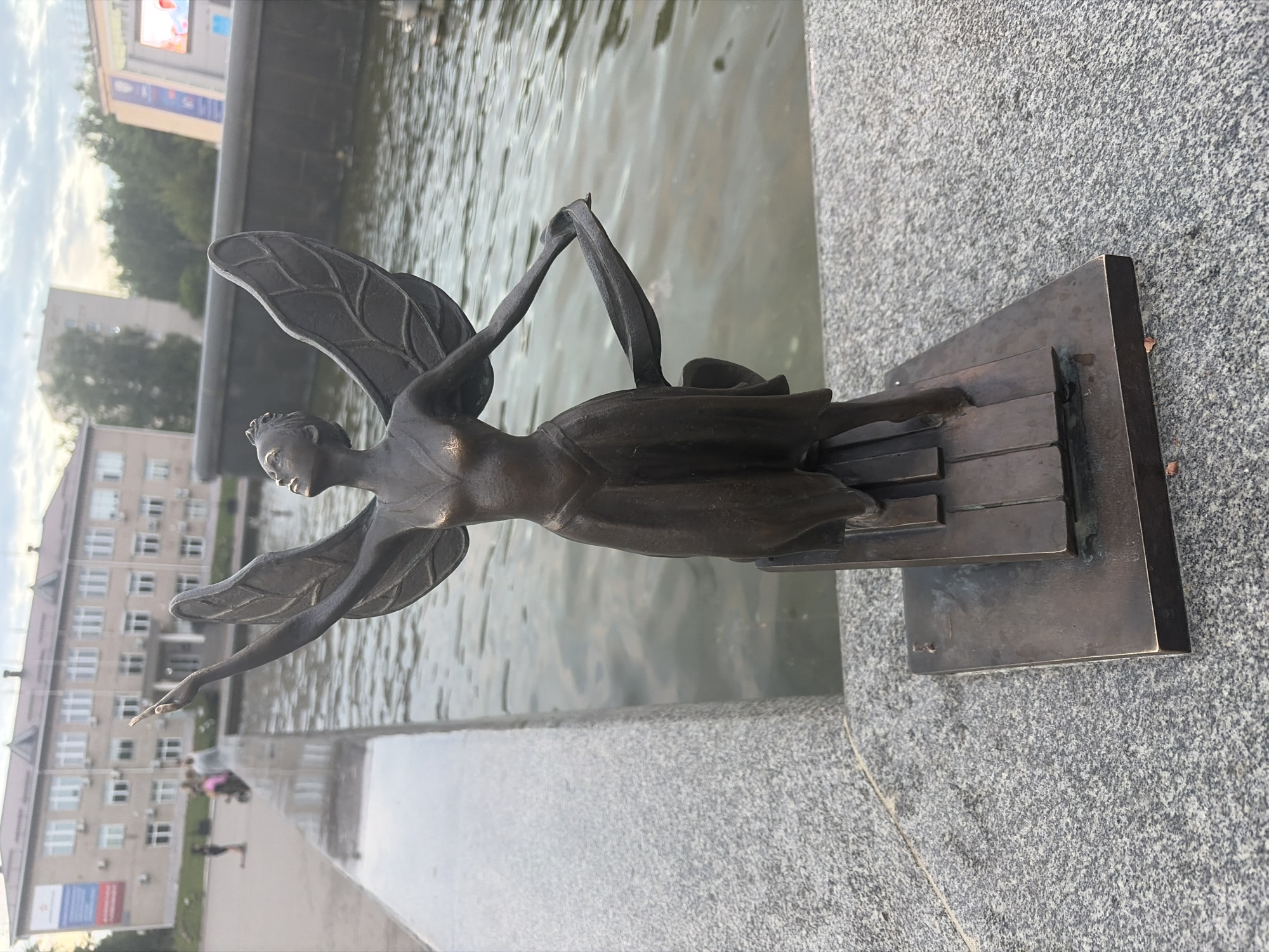 A bronze statue of a winged female figure, resembling a fairy or angel, stands on a granite ledge next to a body of water, likely a fountain or pond. The statue is depicted in a graceful pose, with arms outstretched and wings spread, and is adorned in a flowing dress. The figure stands on a base that resembles piano keys. In the background, a modern building with multiple windows and air conditioning units is visible, along with trees and a cloudy sky. The scene appears to be in an urban setting, possibly a public square or park, identified as being in the city of Chaikovsky, Russia. The lighting suggests it is either early morning or late afternoon.