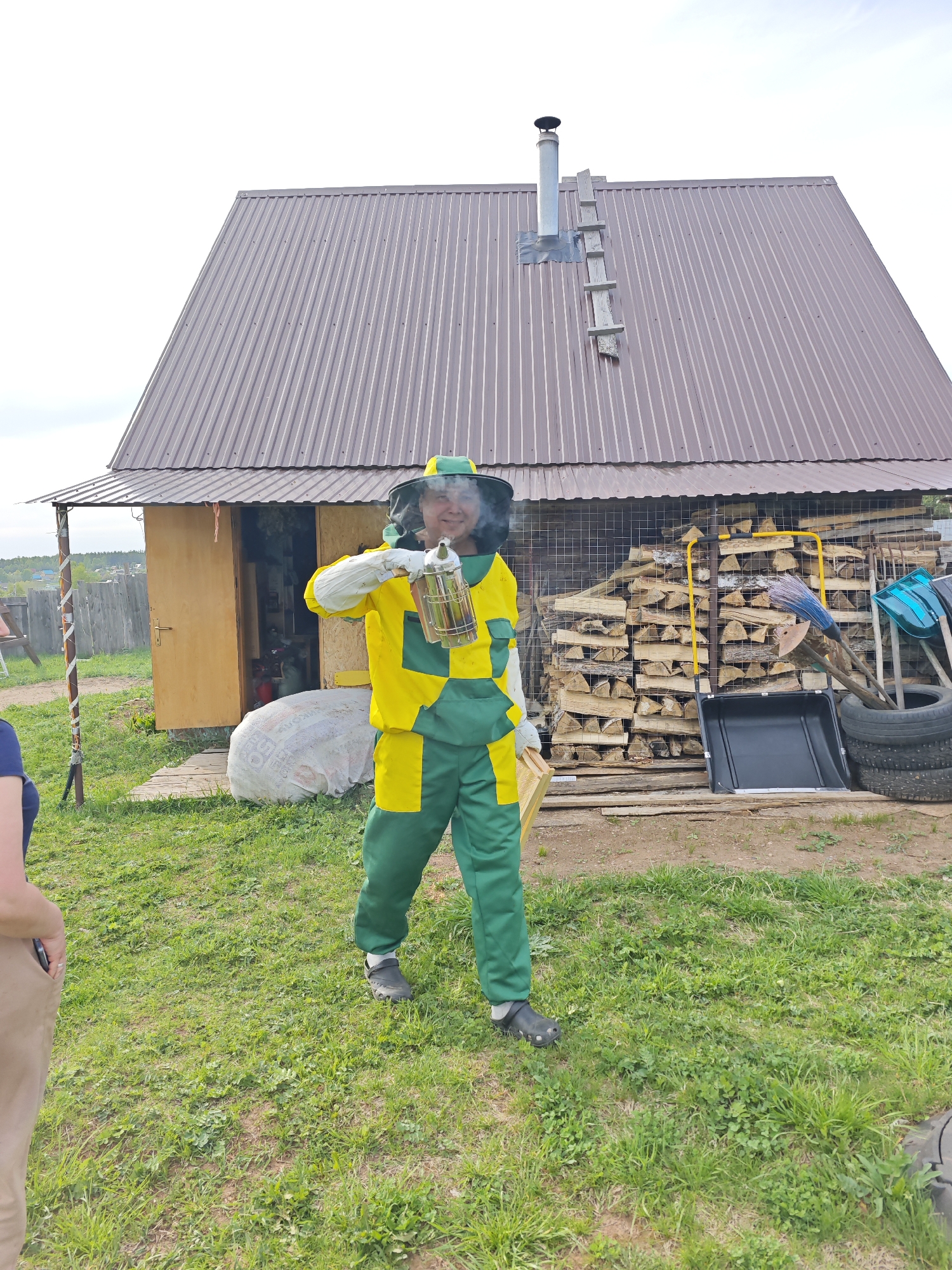 A beekeeper in a yellow and green protective suit and mesh face covering holds a bee smoker in his right hand and a wooden bee frame in his left hand. The beekeeper is standing on green grass in front of a building with a brown corrugated metal roof. The building has a wood frame and a closed door with a window. A white bag sits near the building's entrance. A pile of cut wood sits behind a wire fence. Tires, a yellow wheelbarrow, and blue gardening tools are arranged to the right of the woodpile. The scene is located in Markovo, Russia.