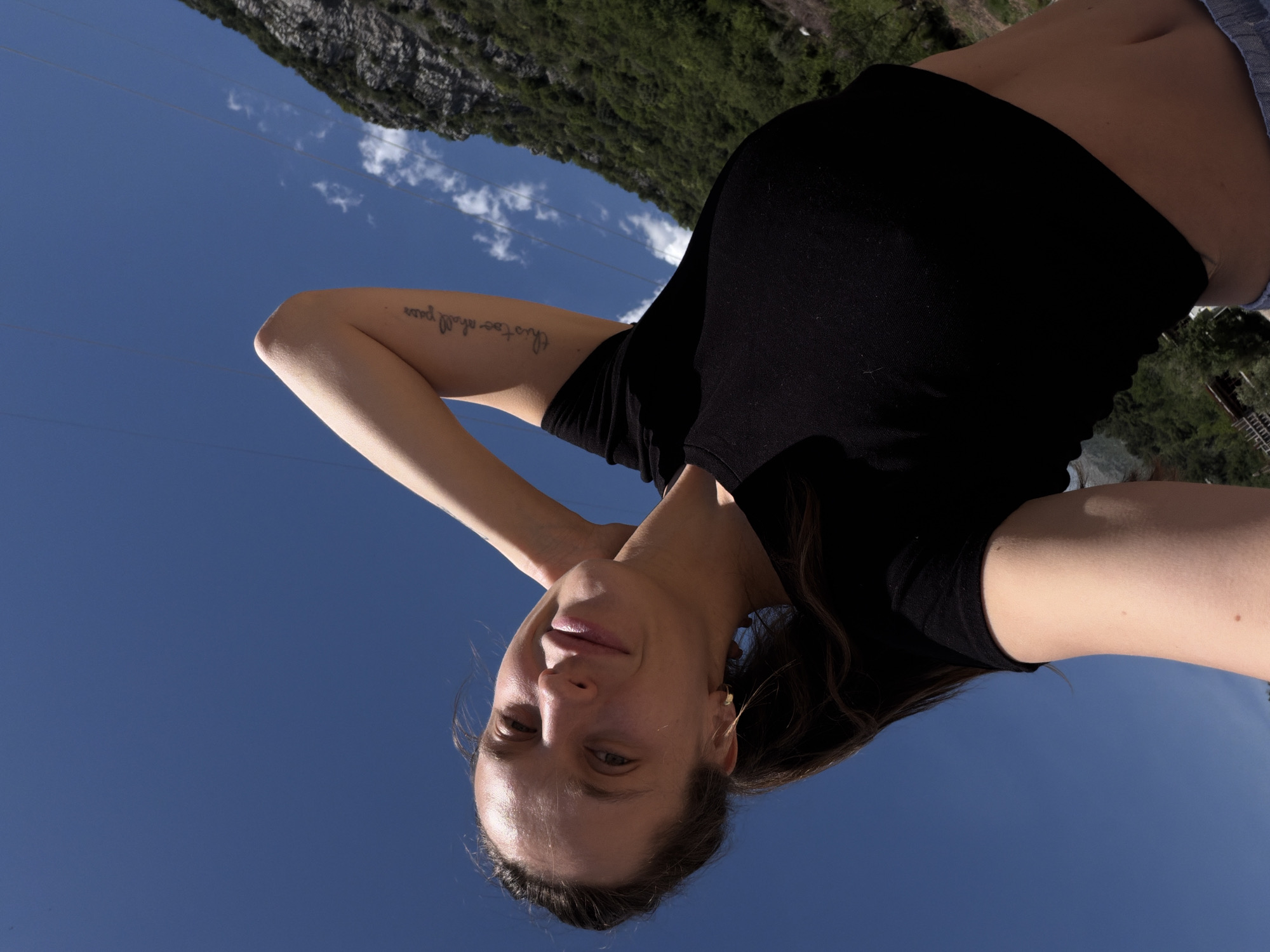 An outdoor, upward-angle shot captures a young woman looking down at the camera, likely a selfie. She has long, dark brown hair and a gentle smile on her face, with a small gold hoop earring visible in her right ear. She is wearing a black top, possibly cropped, revealing her midriff. On her left inner arm, a tattoo reads "this too shall pass." Her right arm is bent with her hand near her neck, while her left arm is visible with the tattoo.
The background features a clear, bright blue sky with scattered wispy white clouds. Thin, dark lines, possibly power lines, traverse the sky. In the upper background, a lush green, rocky hillside or mountain is visible under bright daylight, indicating a sunny day. The scene suggests a relaxed moment in an outdoor setting, consistent with the location of Budva, Montenegro.