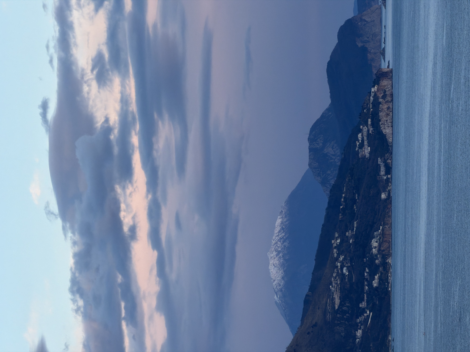 The image shows a landscape with the city of Budva, Montenegro. In the foreground, there is a body of water, likely the Adriatic Sea. Along the coast, a steep, rocky terrain is visible, dotted with white buildings, suggesting residential areas clinging to the hillside. Further back, the view is dominated by mountains that slope into the scene, their details obscured by shadows and the atmosphere. The sky is filled with clouds. The overall impression is one of natural beauty and a scenic coastal settlement.
