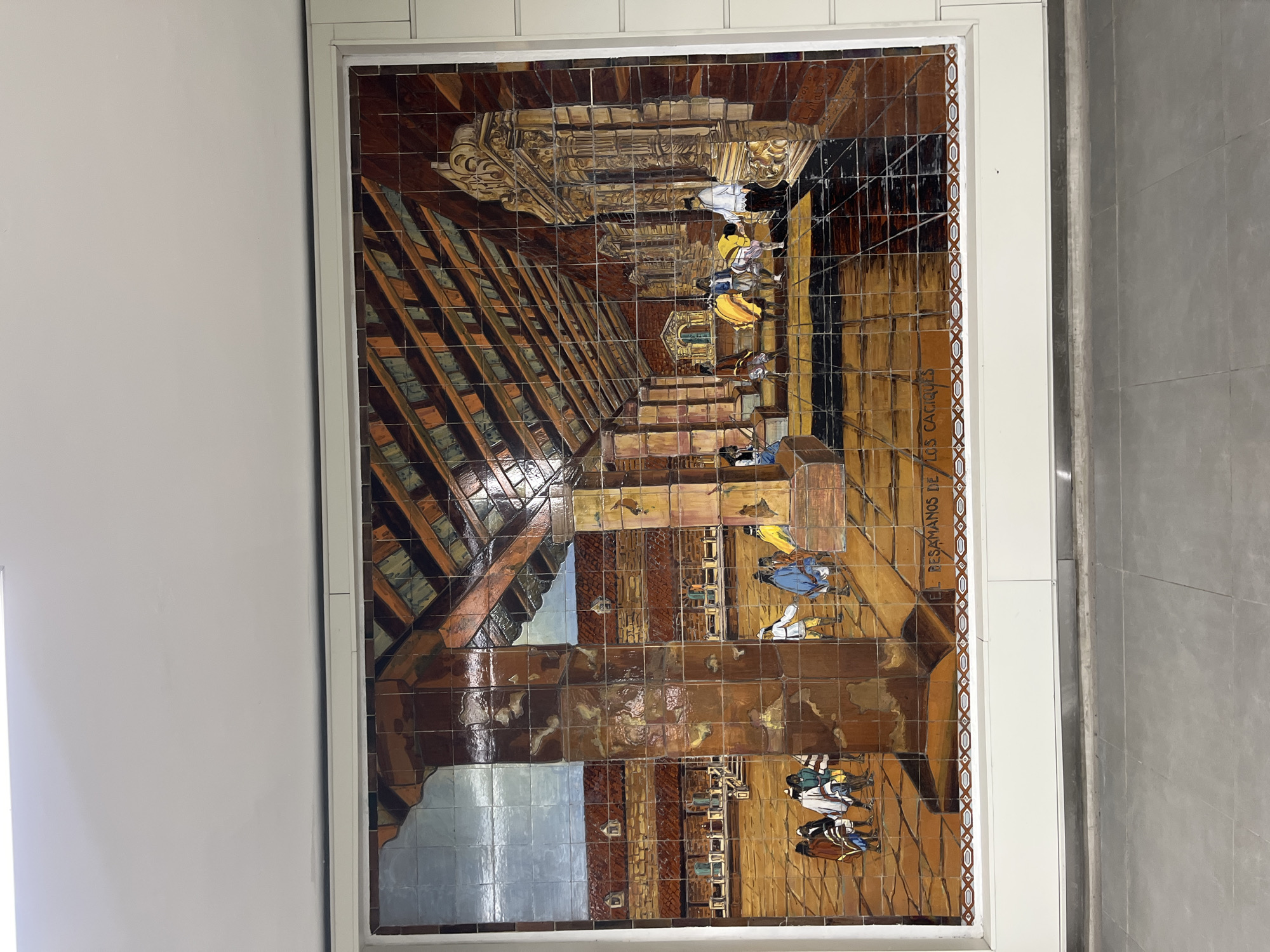 The image depicts a ceramic tile mural, titled "Besamanos de Los Caciques," framed and mounted on a wall. The mural portrays a historical scene with figures dressed in indigenous attire, potentially depicting a meeting or ceremony of chieftains. The scene is set indoors, possibly within a colonial-era building, indicated by the architectural details. The tiles are glazed with a palette of browns, tans, blues, and creams. The text "BESAMANOS DE LOS CACILQUES" appears on the right of the frame. The mural is located in the Perú subway station of Buenos Aires, Argentina.