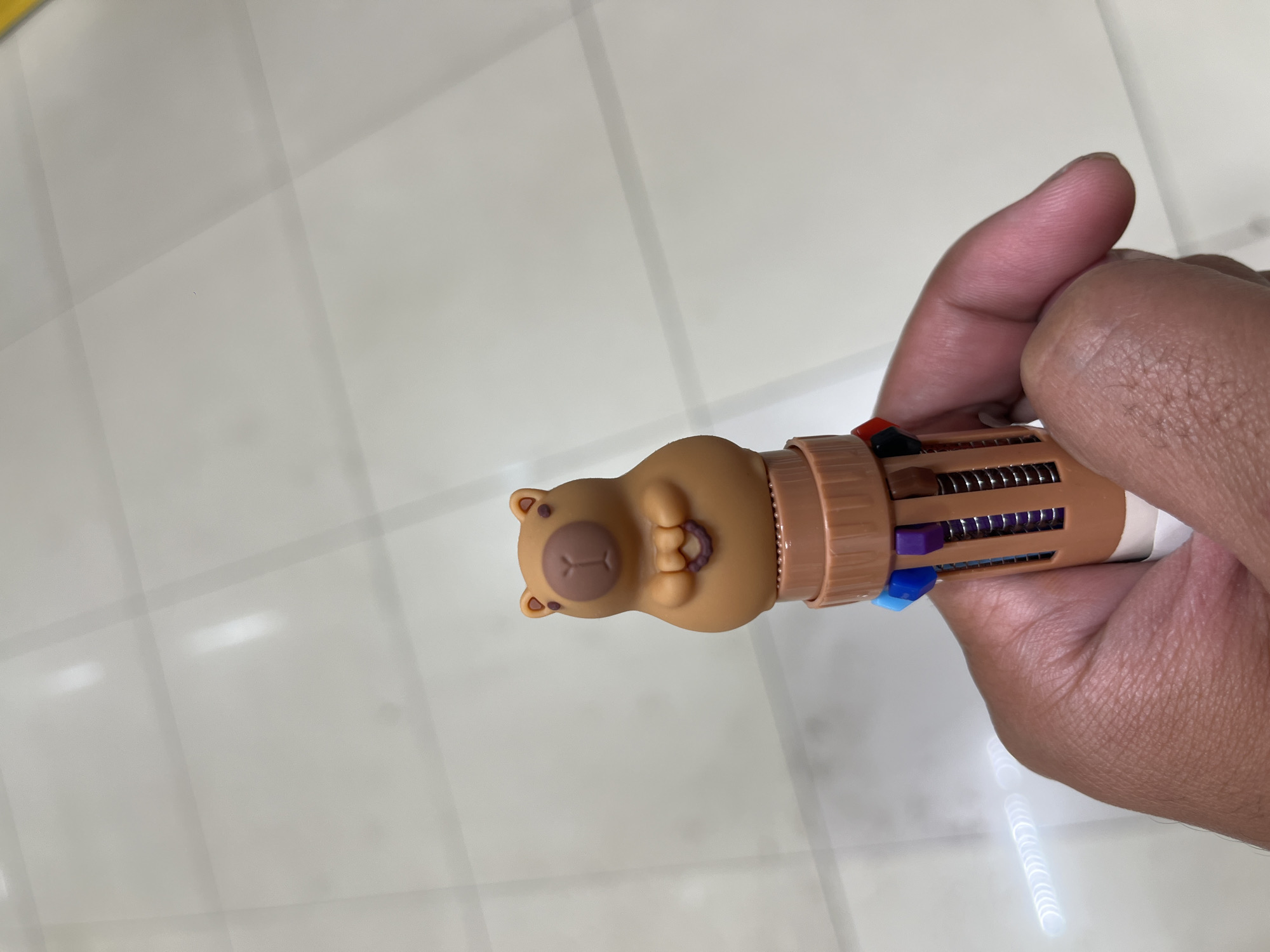 A hand holds a novelty pen with a capybara figurine on top. The pen has multiple colored ink cartridges visible within its barrel. The background is a tiled floor, likely in a public indoor space. The image appears to have been taken in Tangerang, Indonesia.