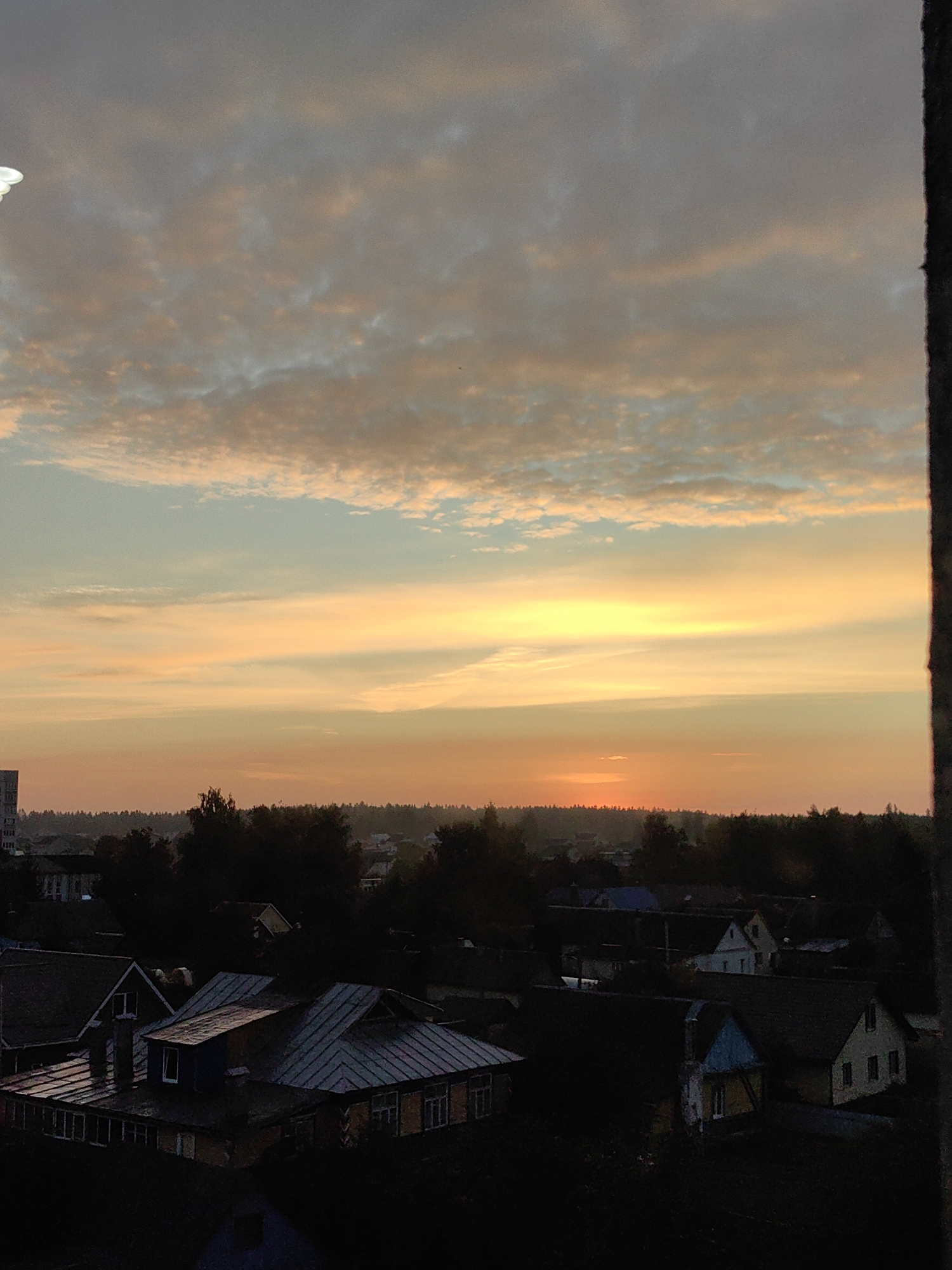 The image shows a sunset over a village in Fanipal, Belarus. The sky is filled with colorful clouds, ranging from pale orange to light blue, with the setting sun casting a warm glow. Below the sky, a dark silhouette of trees and houses forms the landscape. The foreground is dominated by the roofs of several houses, appearing dark and detailed, suggesting a twilight setting. There are no people visible in the image. The overall impression is of a peaceful, rural evening in Belarus.