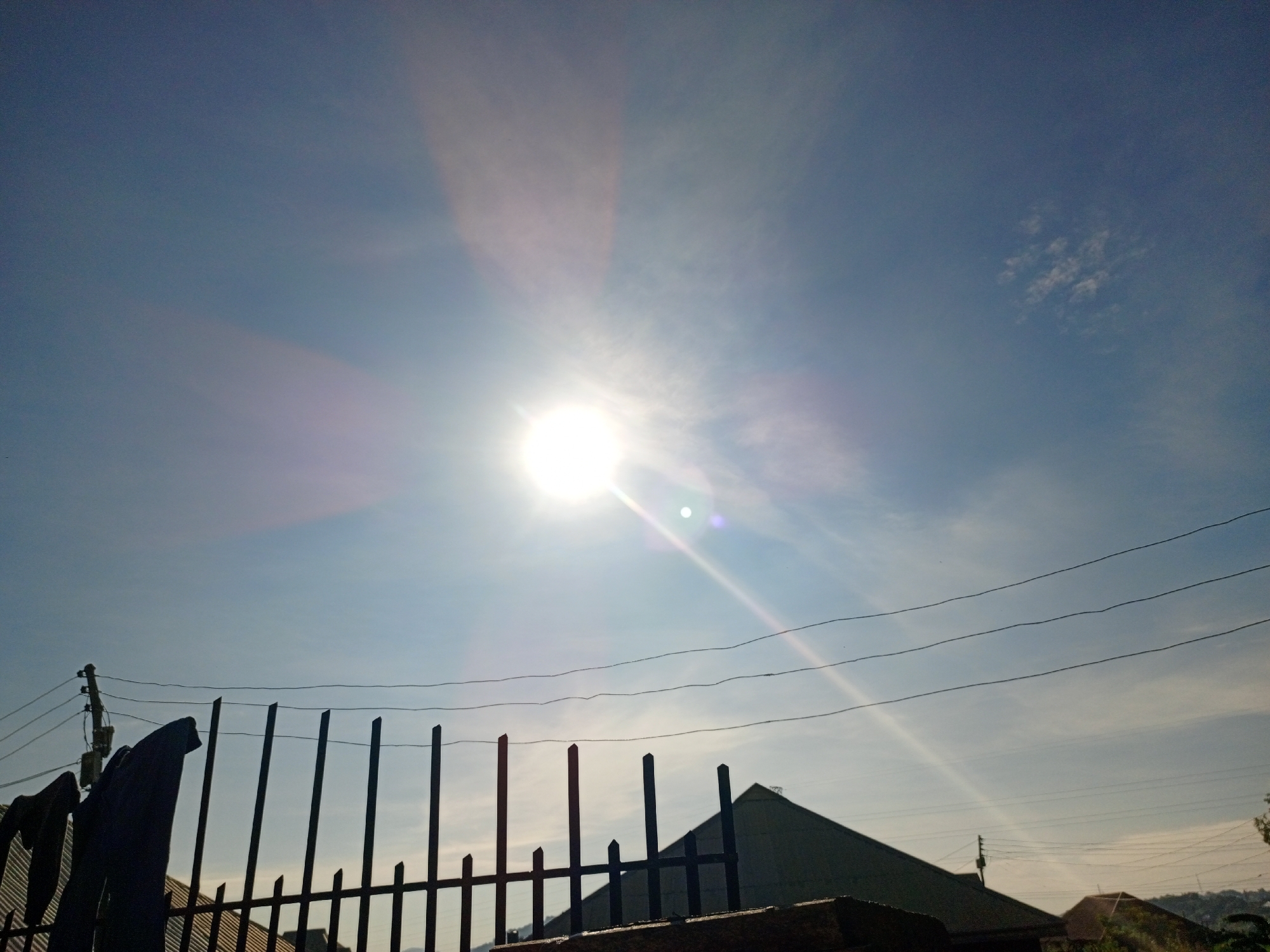 A daytime, exterior, low-angle, wide shot captures a bright sun piercing through a hazy sky. The sun is positioned in the upper center of the frame, casting long rays of light across the blue sky. In the foreground, a dark metal fence with pointed vertical bars runs horizontally across the lower portion of the image. Behind the fence, the dark silhouettes of rooftops are visible. Electrical wires are strung across the sky, supported by a utility pole on the left side of the frame. The overall scene suggests a somewhat overcast day with the sun struggling to break through a layer of haze or light clouds, giving the impression of a warm climate. This scene is located on Rukuba Road, Nigeria.