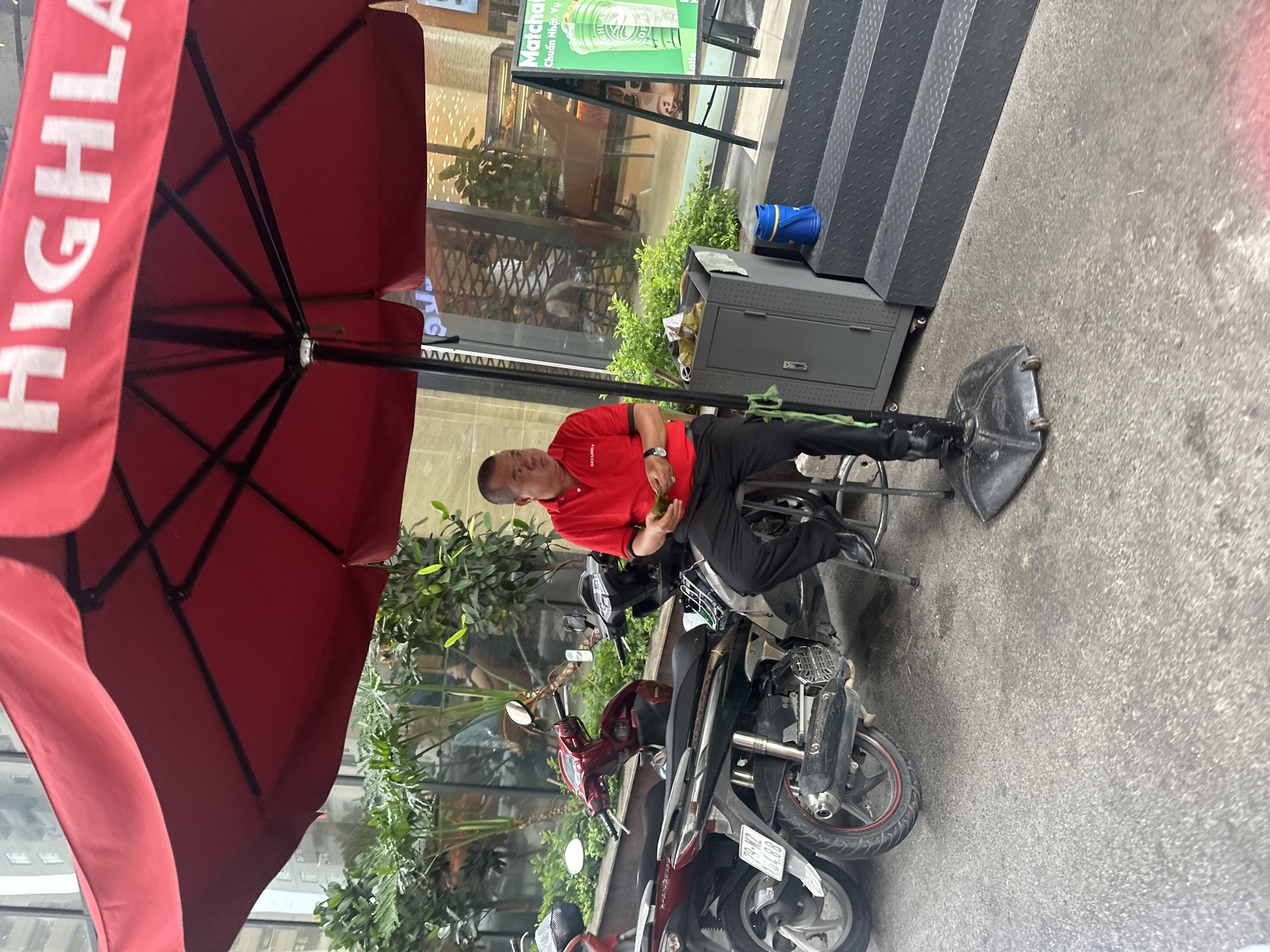 A middle-aged man wearing a red polo shirt with the word "SECURITY" printed on it and black pants is sitting on a stool next to a parked scooter and a set of stairs. He appears to be resting. The scene is outside a building with the sign "Highland" on the red umbrella. The environment suggests an urban setting, likely in or near Bac Nha Trang Ward, Vietnam, given the contextual information provided. The scooter is parked close to the building, and other motorcycles can be seen nearby.