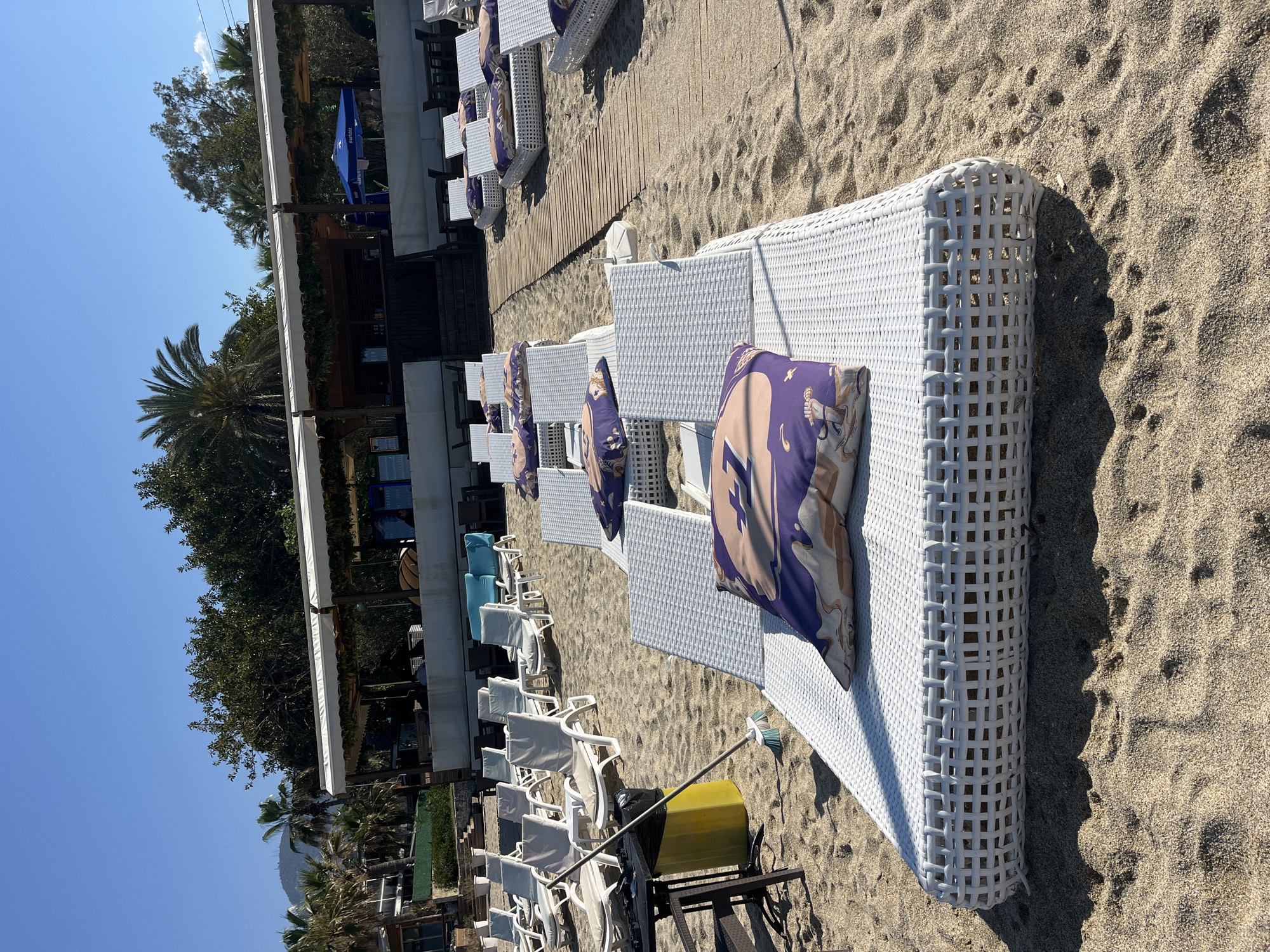 The image shows a beach scene in Alanya, Turkey. Rows of white wicker lounge chairs with purple and tan cushions are lined up on the sand. A wooden boardwalk runs parallel to the chairs. A beachside structure with a white awning and blue umbrellas can be seen in the background. Palm trees and other vegetation are also visible. The sky is clear and blue. A yellow cleaning container on a metal stand, with a broom and a black plastic bag, is visible among the chairs.