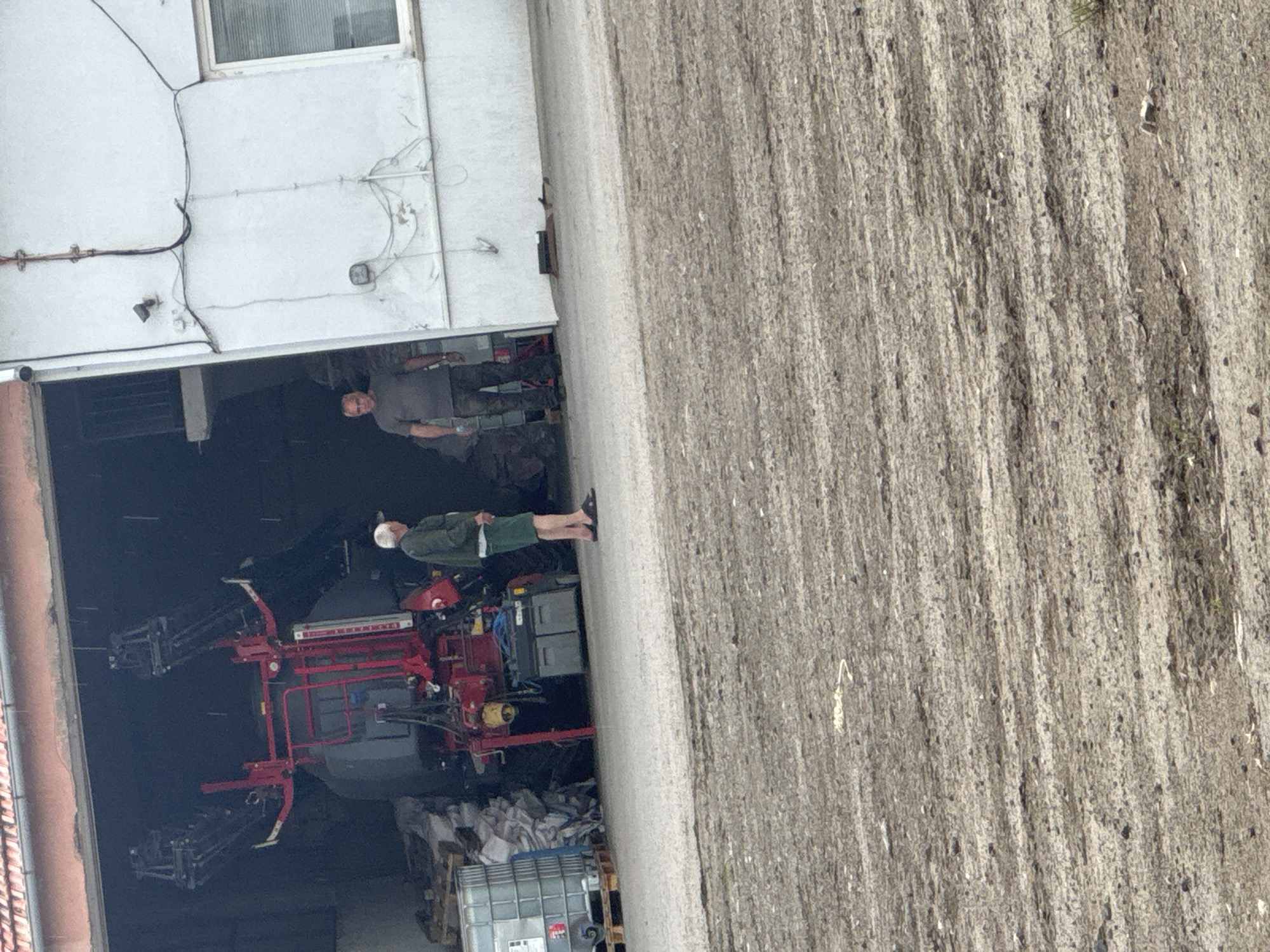 The image depicts a scene in Sarnevo, Bulgaria, showing a white building with a large open garage. Inside the garage, there is an agricultural sprayer attached to a tractor. Two men are present: one is standing outside the garage, wearing a green jacket and light shorts, and the other is inside the garage, wearing a gray shirt and camouflage pants. The area adjacent to the building appears to be a field. A large container sits near the front of the garage.