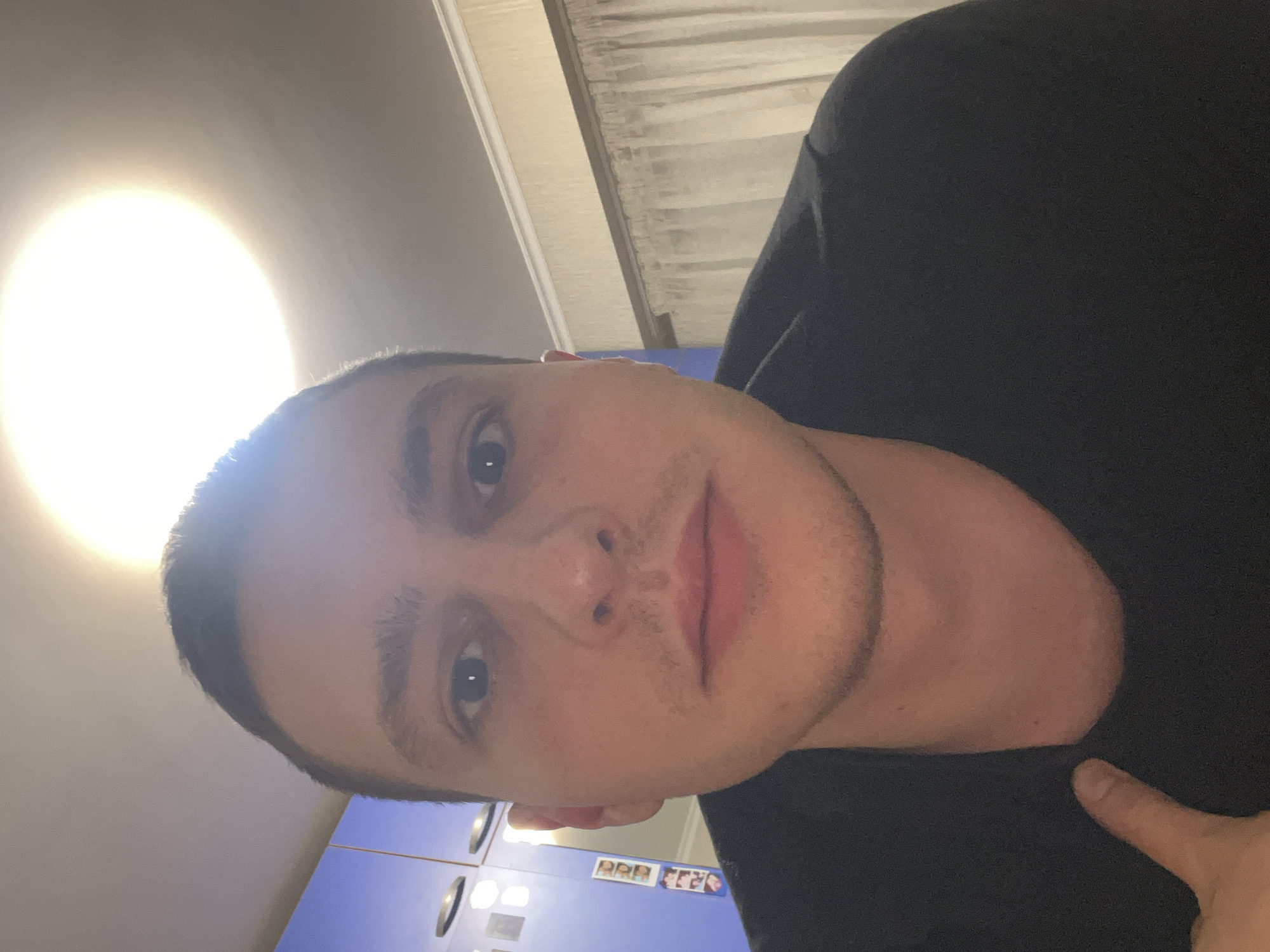 A close-up portrait of a light-skinned male with short hair. He is wearing a dark-colored shirt. The background includes a bright, diffused light source, a textured ceiling, and a blue cabinet with attached photographs. I cannot determine a specific location in Burgas, Bulgaria.