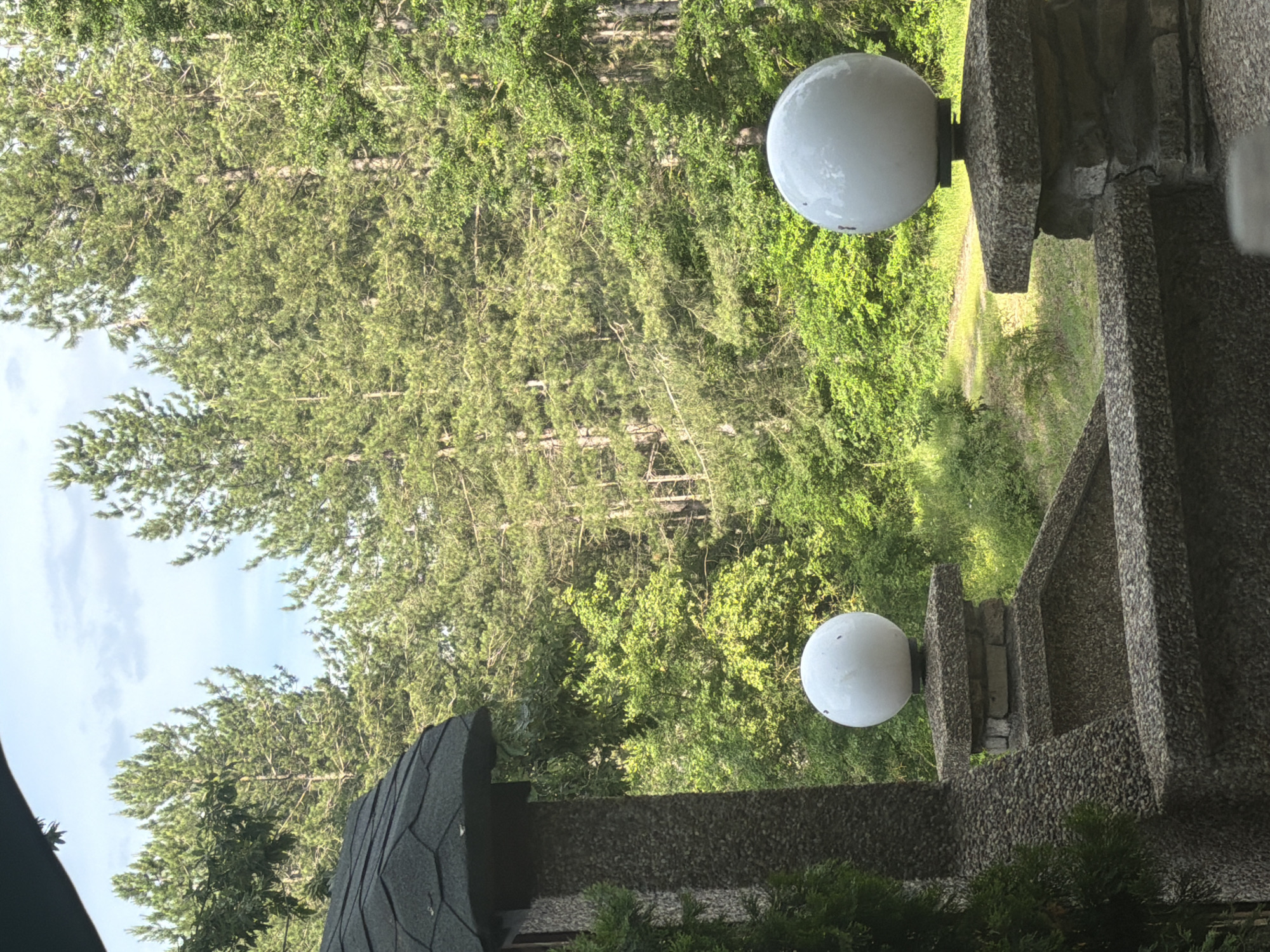 This image captures a serene outdoor scene in Sushina, Bulgaria, characterized by a blend of lush nature and rustic stone architecture, viewed under a partly cloudy sky.
In the midground on the right side of the frame, two prominent spherical, white outdoor light fixtures are visible. These globe lights are mounted on sturdy stone pillars or walls, which exhibit a textured, pebbled concrete or exposed aggregate finish in shades of grey and brown. These stone structures suggest they are part of a railing, fence, or building facade.
The background is dominated by a dense, vibrant forest, primarily composed of tall conifer trees with their characteristic needle-like foliage, interspersed with some lighter green deciduous trees. A narrow dirt path or clearing can be seen winding through the abundant greenery in the mid-right portion of the forest, hinting at trails within the woods.
In the lower-left corner, a dark-colored, shingled roof structure, possibly belonging to a building or gazebo, is partially visible. The sky in the upper-left of the image is partly cloudy, suggesting a daytime scene, likely morning or late afternoon, given the soft, diffused natural light. No people or specific activities are present, contributing to the peaceful and quiet ambiance of the natural setting. The overall impression is one of tranquil nature meeting simple, functional man-made elements.