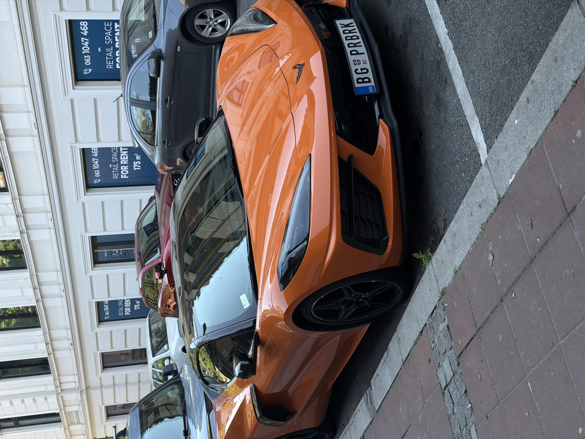 The image shows an orange Chevrolet Corvette parked on a city street in Belgrade, Serbia. The car has a Serbian license plate that reads "BG PRBRK". Behind the Corvette are several other parked vehicles of varying colors. On the left of the frame, the facade of a building is visible with signs advertising "RETAIL SPACE FOR RENT" with the phone number "063 1047 468" and an area of "175 m²". The street surface is a combination of asphalt and red brick paving.