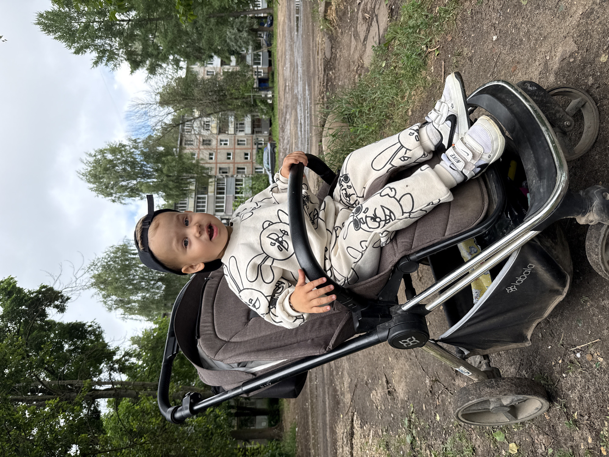 A young child wearing a white sweat suit with a black bunny pattern and a black baseball cap backwards is sitting in a brown stroller. The child's mouth is open, as if speaking or laughing. The stroller has black handles and a black basket underneath. The child is wearing white Nike sneakers with black accents. The stroller is on a dirt path, with grass and trees in the background. In the distance, there are apartment buildings and a cloudy sky.