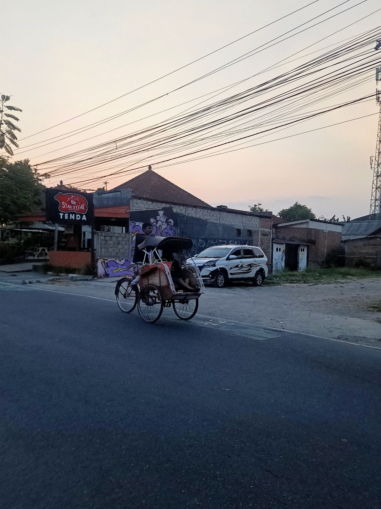 A street-level, eye-level, medium shot captures a three-wheeled bicycle taxi, known locally as a "becak," in motion. The becak is driven by a man, with another person seated behind him. The vehicle is on the right side of a paved road, which occupies the lower half of the frame. In the background, a building with a sign reading "STAR STEAK TENDA" is visible, suggesting a commercial establishment. Adjacent to it are other structures, including what appears to be a small white building with a door and a white SUV parked in front of a row of buildings. Overhead, a dense network of electrical wires stretches across the sky, which is a pale orange, indicative of either sunrise or sunset. A cell tower is visible on the far right of the frame. The overall setting suggests an urban or semi-urban environment in Klaten, Indonesia, during a period of low light.