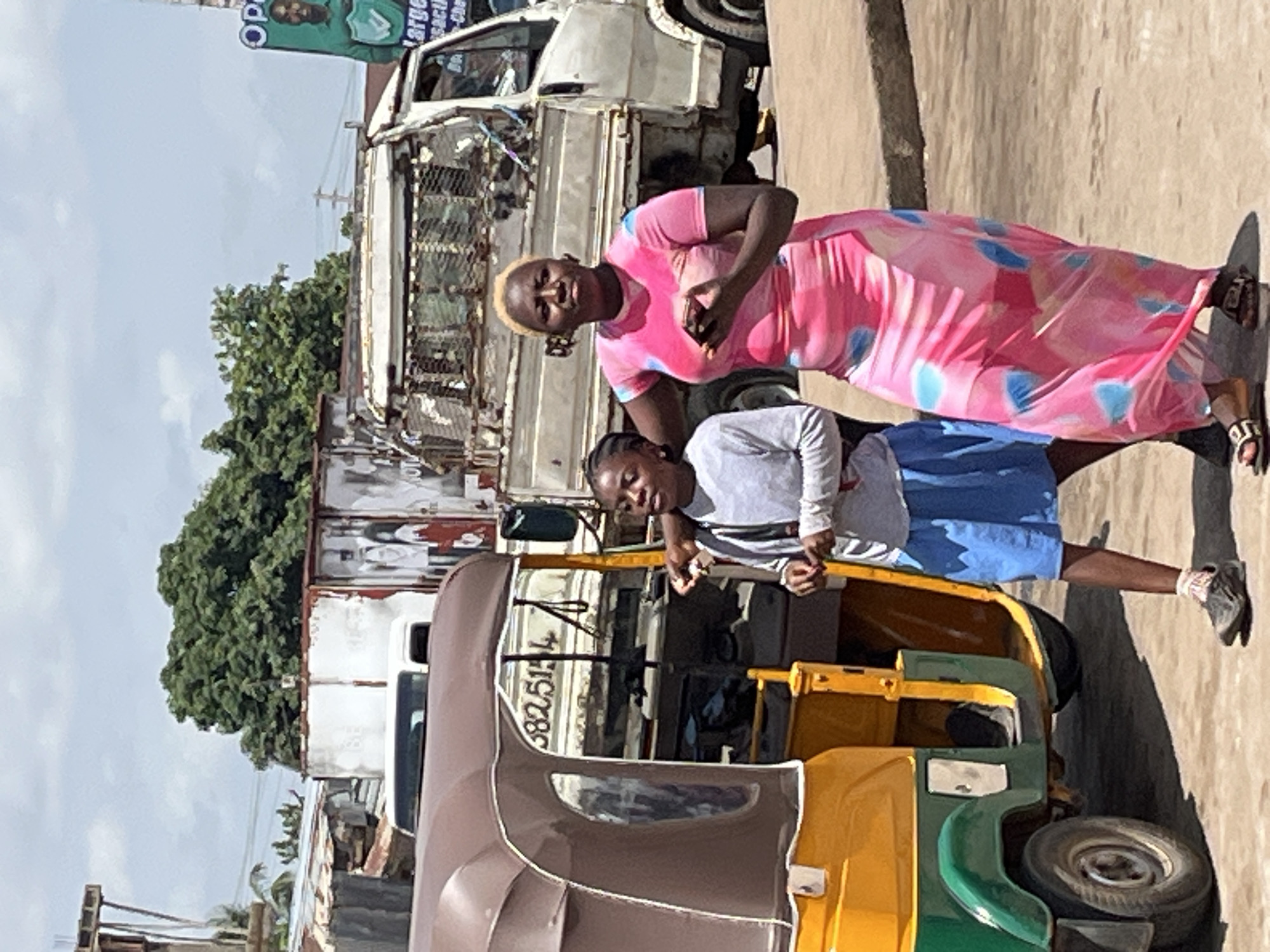 The image shows two young females standing next to a yellow and green auto-rickshaw. The female on the left wears a light grey long-sleeved shirt and blue shorts. The female on the right wears a long pink dress with blue and white tie-dye patterns. She is holding a smartphone in her right hand. In the background, there is a white truck with a metal cage, a white shipping container, and various buildings and vegetation. The ground appears to be a dirt road. The scene is consistent with an outdoor market or street environment in Gura Top, Nigeria, characterized by the presence of auto-rickshaws, trucks, and open-air settings.