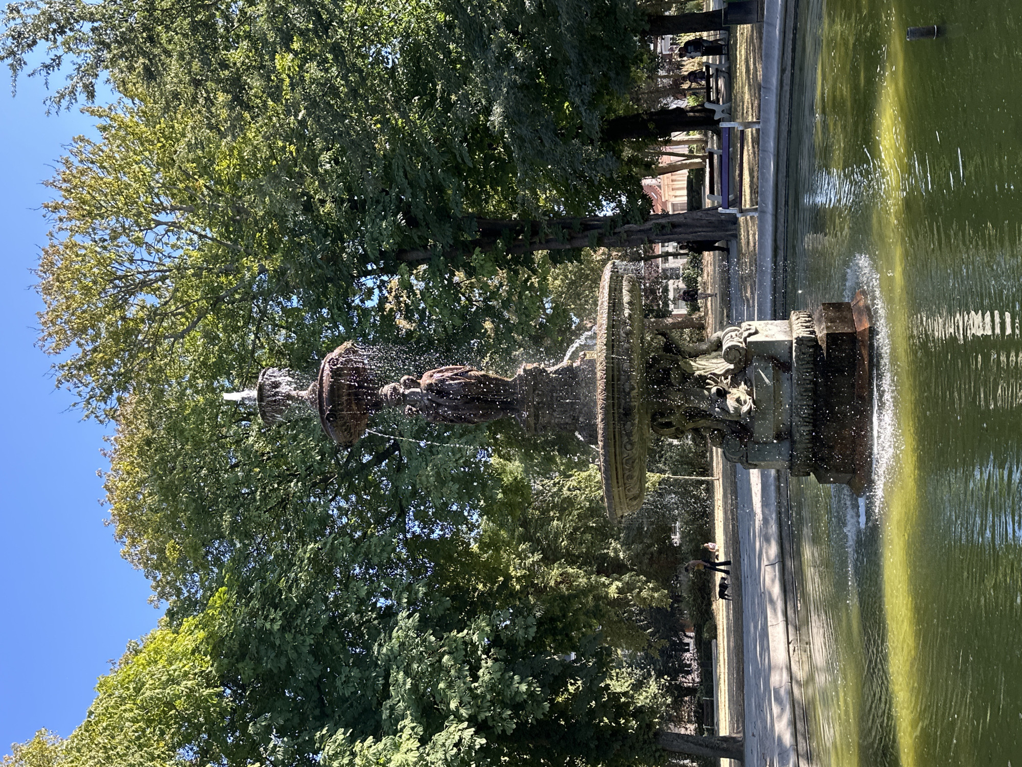 The image shows a fountain in a park. The fountain is ornate and has multiple tiers with water flowing from the top. A statue of a person is visible on the fountain. The fountain is surrounded by a pool of green water. In the background, there are many trees with lush green leaves. Several people are scattered throughout the park, some walking and some sitting on benches. A dog is also present, being walked by one of the people. The sky is clear and blue, indicating a sunny day. The overall scene depicts a peaceful day in a city park. The location is identified as Calais, France.