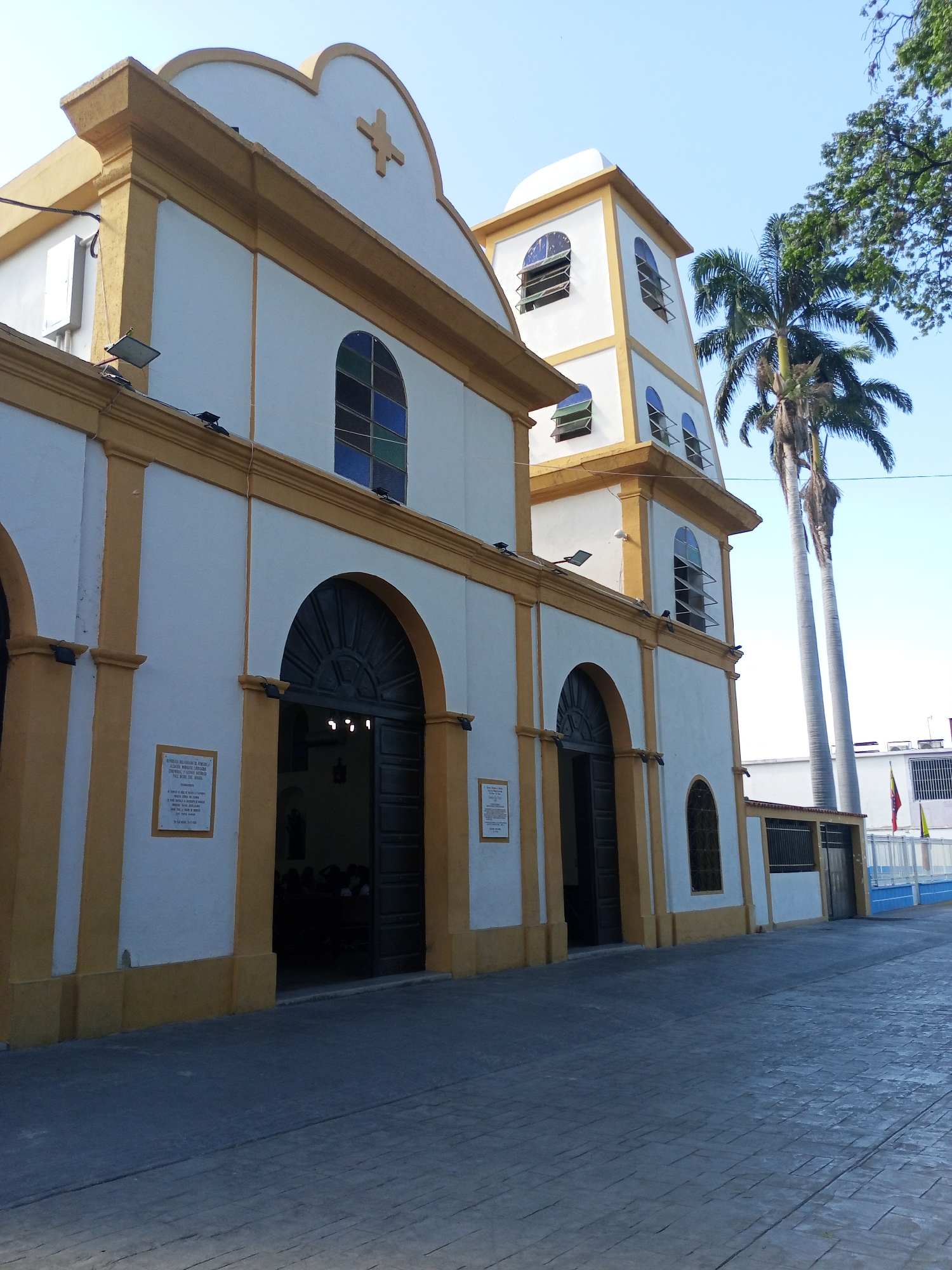 The image depicts the Iglesia San Luis Rey church located in Palo Negro, Venezuela. The church's facade is primarily white with yellow accents outlining architectural features like columns, arches, and the roofline. It has two prominent arched entrances and a multi-tiered bell tower. The sky is a clear, light blue. Palm trees stand to the right of the church. A paved, grey stone walkway leads to the entrance. A plaque is mounted on the wall next to the left entrance. People are inside the church. A Venezuelan flag is visible near the outer fence, along with blue paint along the curb. The image seems to be taken during the daytime.