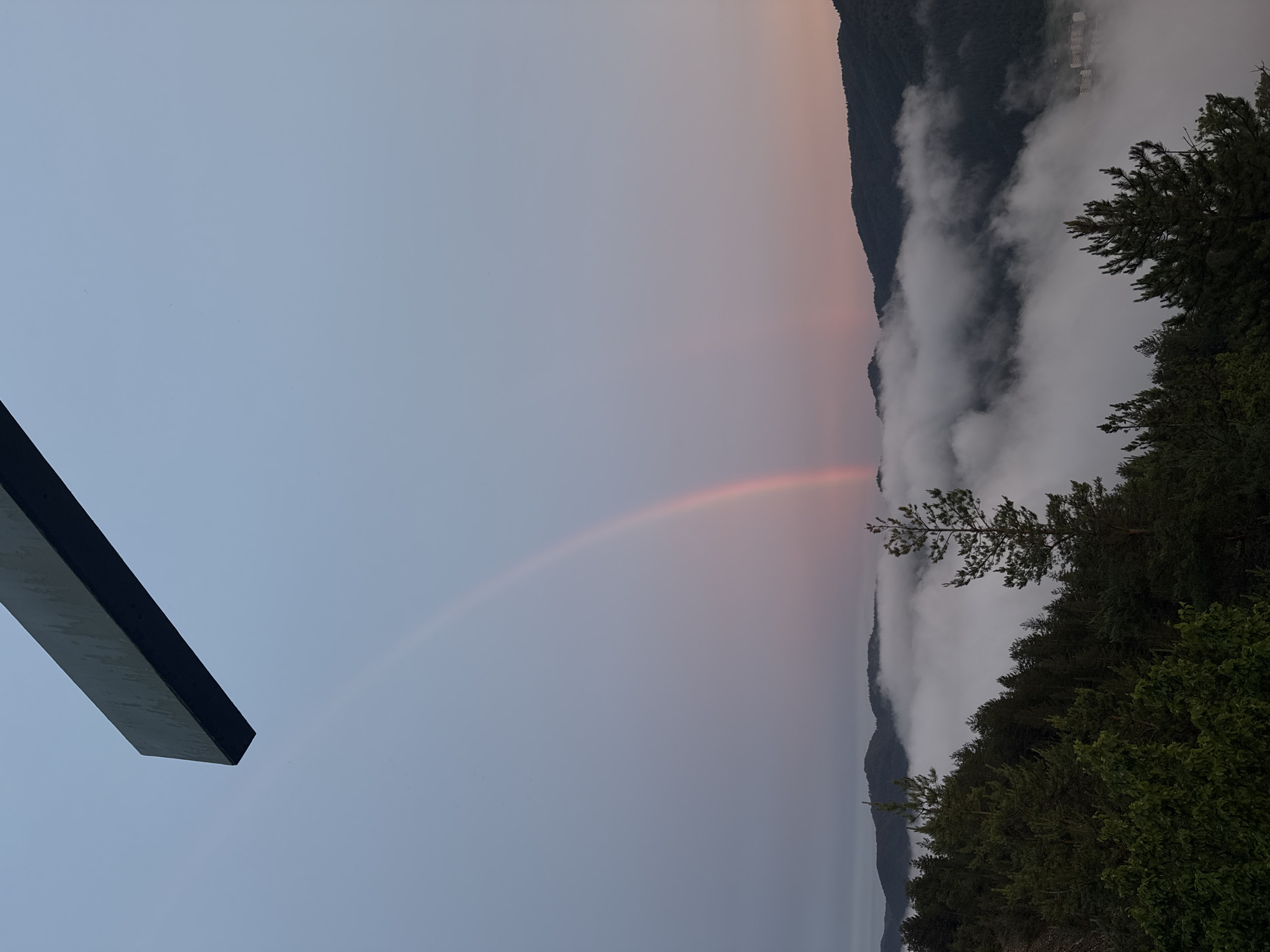 The image shows a natural landscape, likely taken from an elevated viewpoint, in Strazha, Bulgaria. The scene is dominated by a vast expanse of sky and a band of soft, pinkish clouds tinged with the last light of day, suggesting either dawn or dusk. A faint rainbow arcs across the sky, indicating recent rainfall or atmospheric moisture.
Below the sky, a dense layer of white clouds partially obscures a mountainous terrain. The mountains appear as dark, silhouetted forms rising from the cloud cover. The upper slopes of these mountains are covered in dark green forests. In the foreground, on the right side of the image, the dark green foliage of trees, likely conifers and deciduous shrubs, is visible, suggesting a wooded area at the viewpoint.
On the left side of the image, a large, dark, rectangular object juts into the frame from the bottom left corner. Its surface appears to be dark and possibly metallic or stone, with a subtle texture indicating it might be part of a structure, a sculpture, or a viewing platform.
The overall mood is serene and slightly mystical, with the soft colors of the sky, the ethereal clouds, and the subtle rainbow creating a sense of peace and wonder. The time of day is likely early morning or late evening due to the soft lighting and the presence of the rainbow and the atmospheric haze. There is no visible text in the image.