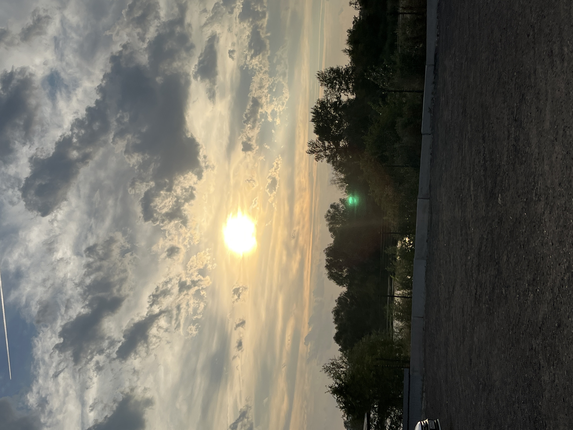 The image depicts a scenic sunset over a body of water, framed by a line of trees. The sky is filled with dramatic, scattered clouds, ranging from dark gray to lighter, wispy formations. The sun, a bright orb, is positioned in the upper center of the frame, casting a warm, golden light that illuminates the clouds and reflects on the water. A faint contrail from an aircraft is visible in the upper left corner of the sky.
In the foreground, a dark, gravelly area suggests a parking lot or undeveloped land. A small portion of a white car is visible on the far left edge of the image, indicating a potential parking area. Beyond the gravel, a low concrete barrier runs horizontally across the scene, separating the foreground from the natural landscape.
The middle ground is dominated by a dense line of trees, their foliage dark and lush, with some variations in height and density. Behind the trees, a body of water, possibly a lake or river, is visible. The surface of the water is calm, with subtle reflections of the sky and surrounding vegetation. A green lens flare is noticeable on the left side of the water, likely caused by the setting sun. A dark metal fence runs along the edge of the water, partially obscured by the trees.
The overall impression is one of natural beauty at dusk, with elements of human presence in the form of the car and the barrier. The location is identified as Shuмейka, Russia, suggesting a rural or semi-rural setting with access to natural water features.