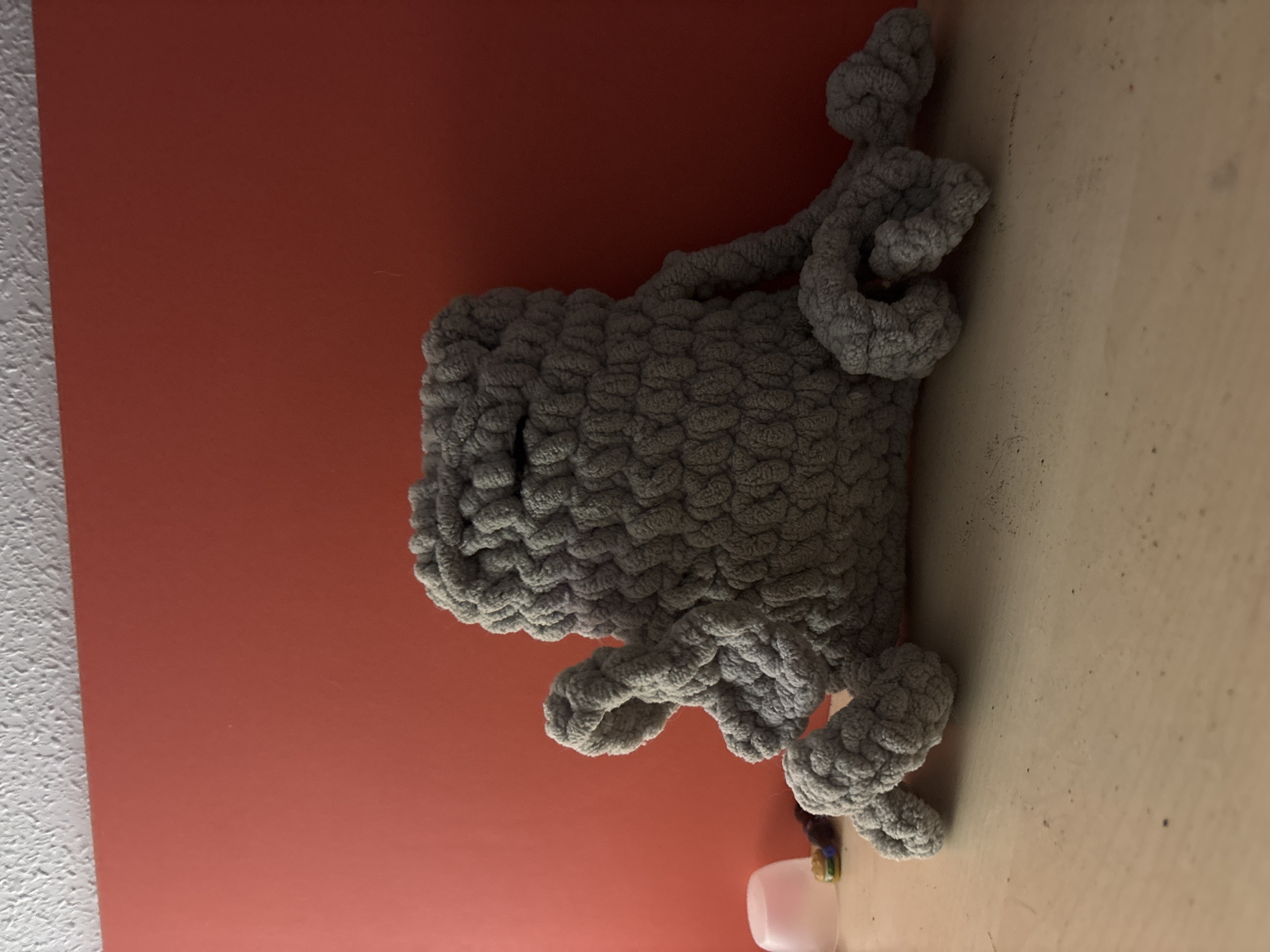 A chunky, grey knitted toy is placed on a textured surface against a coral-colored wall. The toy resembles an animal, possibly a dinosaur or a mythical creature, with a bulbous body and multiple limbs that are curled and looped. The knitting technique creates a distinct, textured pattern across the entire toy. The background wall is a solid, muted coral or terracotta color. To the left of the wall, there is a strip of white, textured material, which might be a ceiling or another wall surface. The surface the toy rests on is a light beige or cream color and appears to be a table or shelf, with some subtle dark specks and smudges visible. In the lower right corner, a portion of a small, white object, possibly a container or lid, is visible. The lighting suggests an indoor setting, with shadows cast by the toy indicating a light source from the right. No people are visible in the image. The scene is static, with the toy simply positioned in the frame. There is no visible text. The location context of Orihuela, Spain, does not offer any specific visual cues in this image.