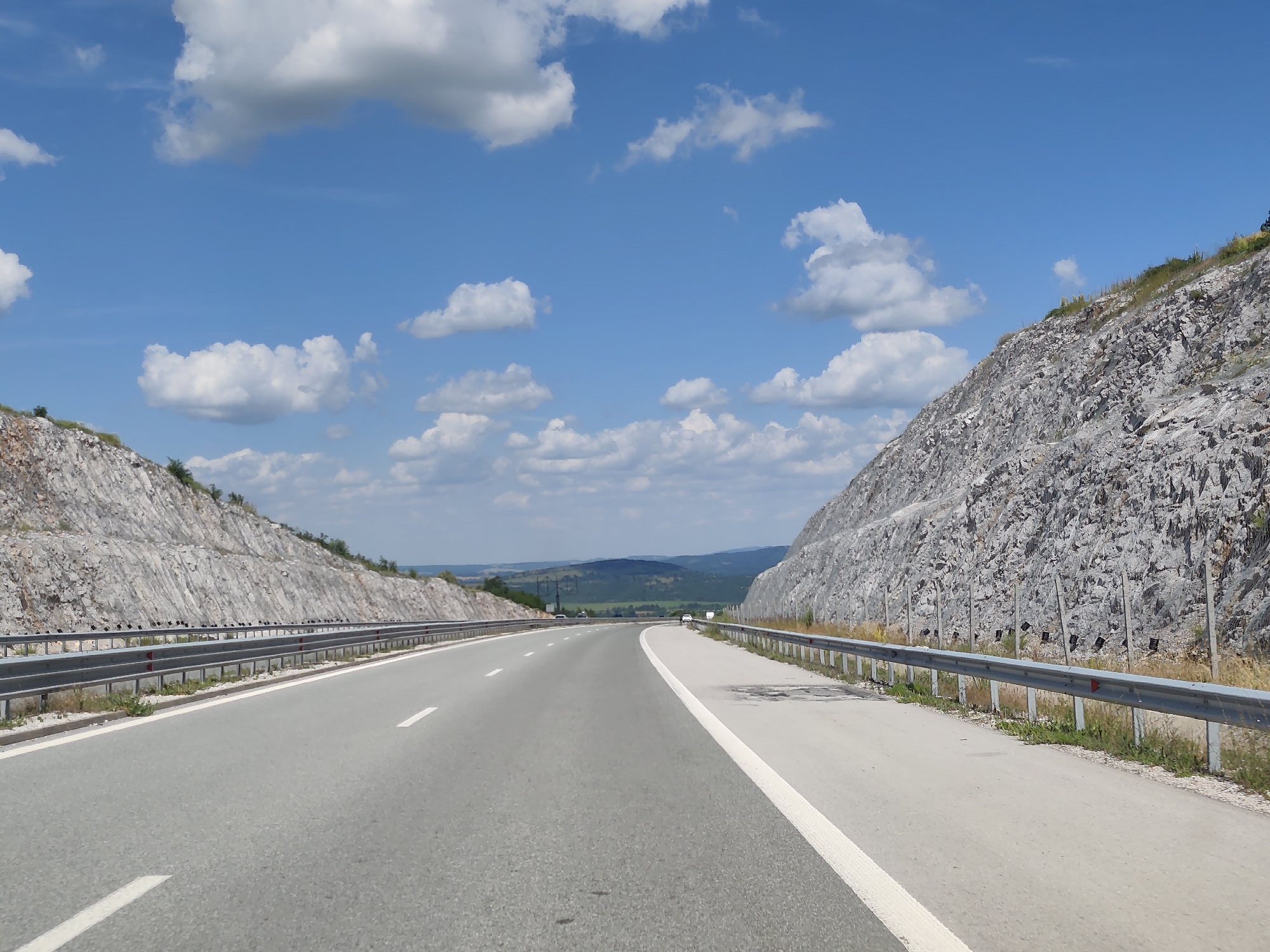The image captures a highway cutting through a mountainous landscape near Yablanitsa, Bulgaria. The road is paved and has clear white lane markings. On either side of the road, steep, rocky hillsides rise dramatically, appearing to have been cut to make way for the highway. A silver guardrail runs along the edge of the road on both sides. In the distance, rolling hills and mountains are visible under a bright blue sky dotted with white, fluffy clouds. A few scattered vehicles are present on the highway, moving in the distance. The environment appears to be arid or semi-arid, with sparse vegetation growing on the slopes and along the roadside. The overall scene suggests a scenic drive through a geologically interesting area.
