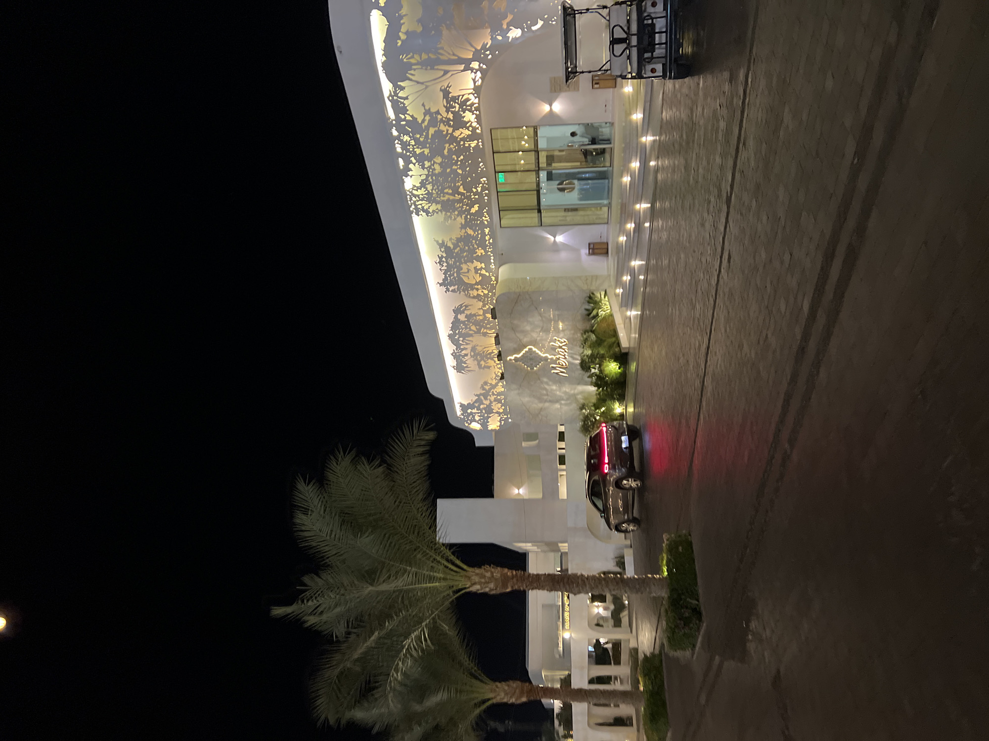 A dark, clear night sky with a full moon is visible above a modern, white building with large palm trees in the foreground. The building features intricate, decorative white cutouts of trees and leaves on its facade, illuminated by warm lights. A stone walkway with scattered lights leads to the entrance, which has glass doors. A dark-colored SUV with illuminated taillights is parked on the right side of the walkway. A white golf cart is parked in front of the building to the right. The ground is paved with cobblestones. The scene is likely from the resort area of Sharm El Sheikh, Egypt, given the presence of palm trees and the resort-like architecture. The overall impression is one of an upscale, tranquil evening setting.