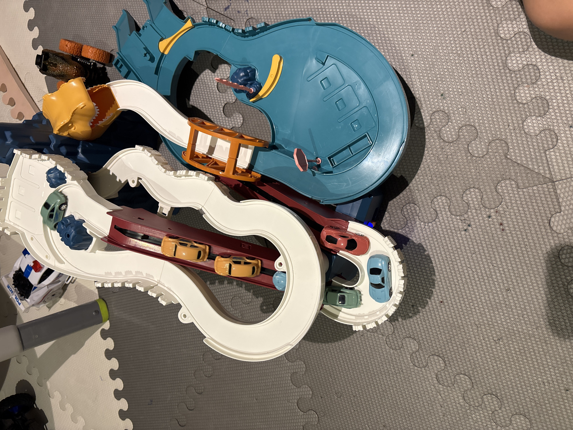 The image shows a toy car race track with several toy cars on it. The race track is made of white and blue plastic and has a dinosaur head at one end. There are several toy cars on the track, including a yellow car, a red car, a blue car, and a green car. There is also a police car in the background. The race track is set up on a grey puzzle mat. The scene appears to be indoors. There are no people visible in the image. The image does not contain any information that would indicate its location in Dubai, United Arab Emirates.