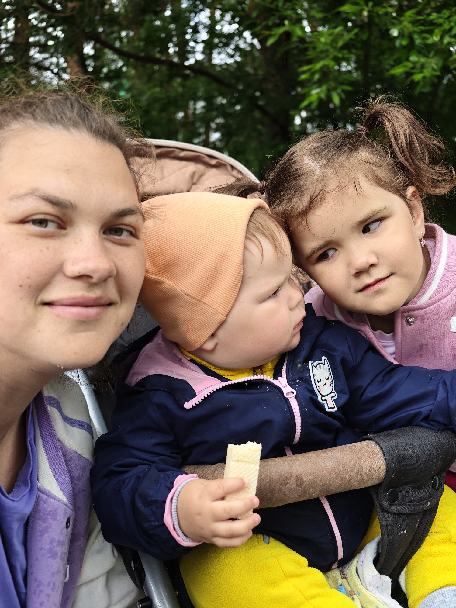 A close-up selfie captures an adult woman with two young children, likely outdoors in a park or garden setting, with the given context of Chaikovsky, Russia. The woman, positioned on the left, has brown hair, a light complexion with freckles, and a gentle smile. She is wearing a purple and white striped garment beneath a purple jacket.
To her right, a baby or toddler sits, wearing an orange beanie, a dark blue jacket with a pink zipper and an embroidered white cartoon cat patch on the chest, and bright yellow pants. The baby is holding a piece of waffle in their right hand, appearing focused on it. Next to the baby, on the right side of the frame, is an older child, a girl with brown hair styled into two pigtails. She wears a pink varsity-style jacket and a small star-shaped earring is visible on her right ear. Her gaze is directed towards the baby with a thoughtful or curious expression.
The children are seated in what appears to be a stroller or similar carrier, with a padded armrest or bar visible in front of the baby. The background is a soft, out-of-focus blur of green foliage, suggesting a natural outdoor environment during daytime. The scene conveys a calm, affectionate family moment.