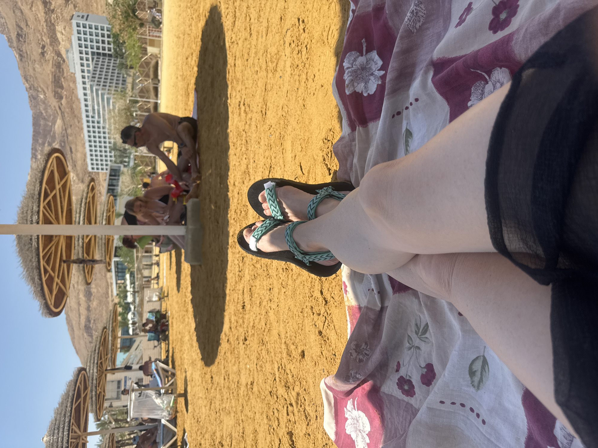 The image shows a view from a reclining position on a beach. The location is likely in the Tamar Regional Council, Israel, given the presence of the Dead Sea. A person is lying on a patterned blanket on the sand, with crossed legs and feet in teal and black sandals. In the background are straw umbrellas, other people relaxing on the beach, a white building, and a rocky mountain range. The scene is bathed in sunlight.