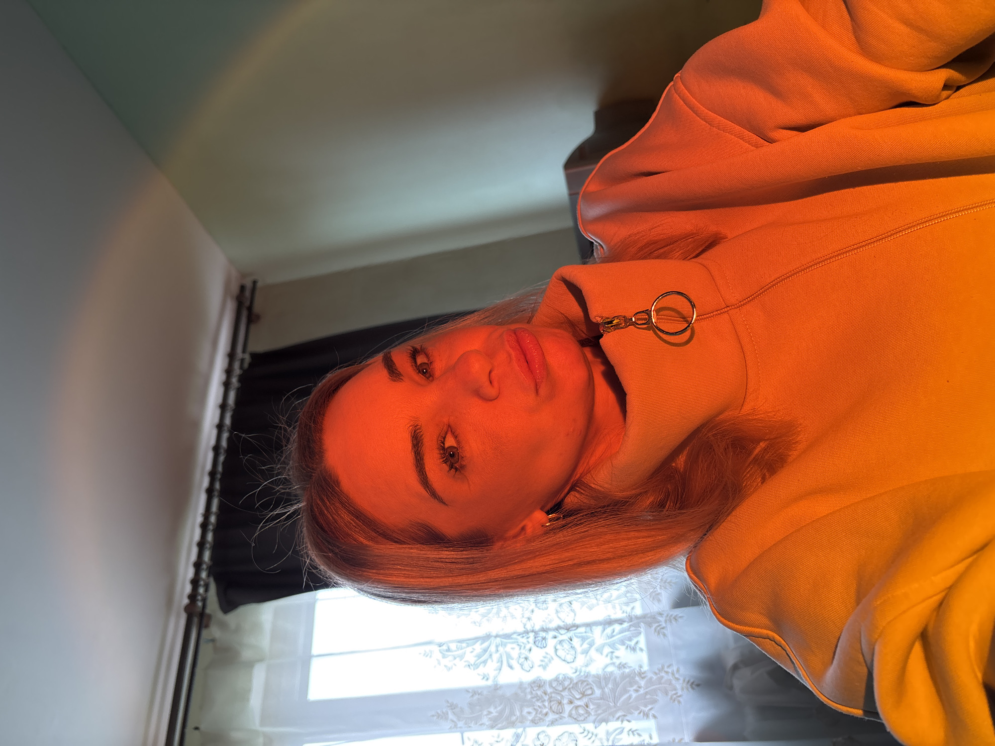 A selfie of a light-skinned woman with blonde hair and green eyes, wearing a tan, zip-up sweatshirt with a circular zipper pull. The background shows a white wall, a window with a patterned sheer curtain, and a dark curtain partially visible. The lighting is warm and somewhat orange. The location is Unknown, Unknown.